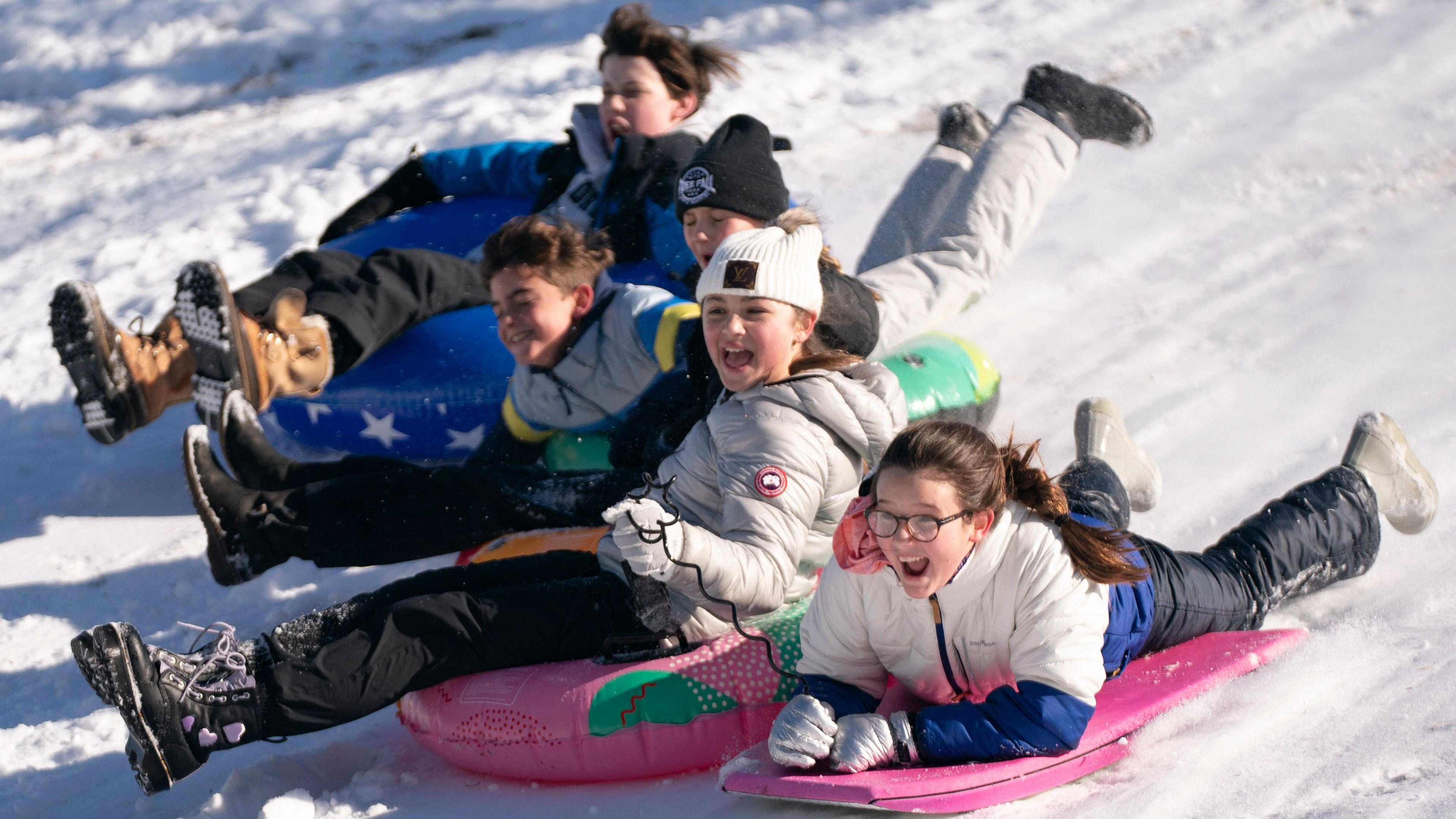 Fünf Kinder in Winterkleidung rodeln auf bunten Schwimmringen einen Schneehügel hinunter. Ein Mädchen mit Brille liegt mit auf dem Bauch auf einem pinken Surfbrett und 