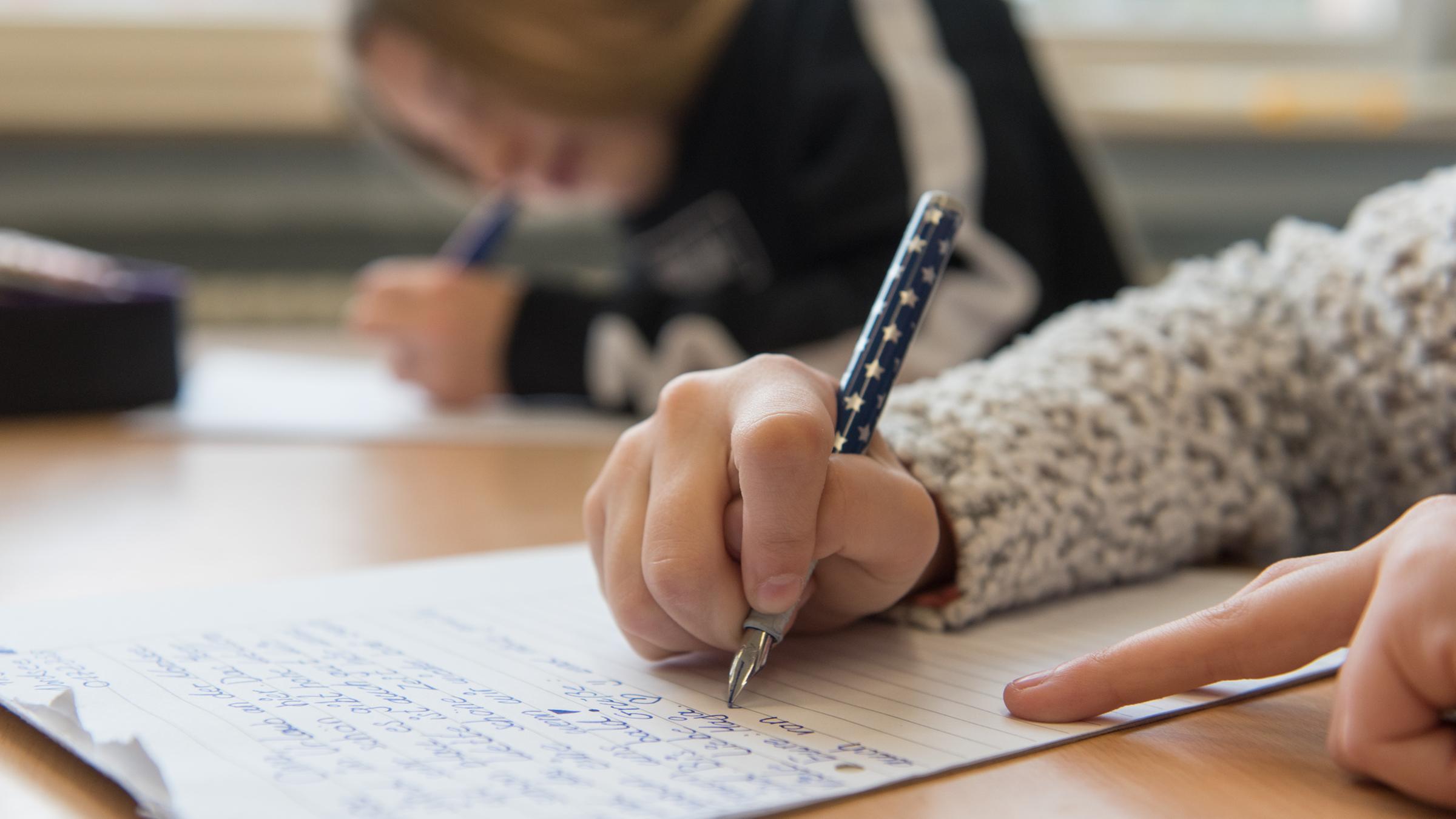 Die Hand einer Schülerin schreibt handschriftlich einen Text auf einen Schreibblock im Schulunterricht.