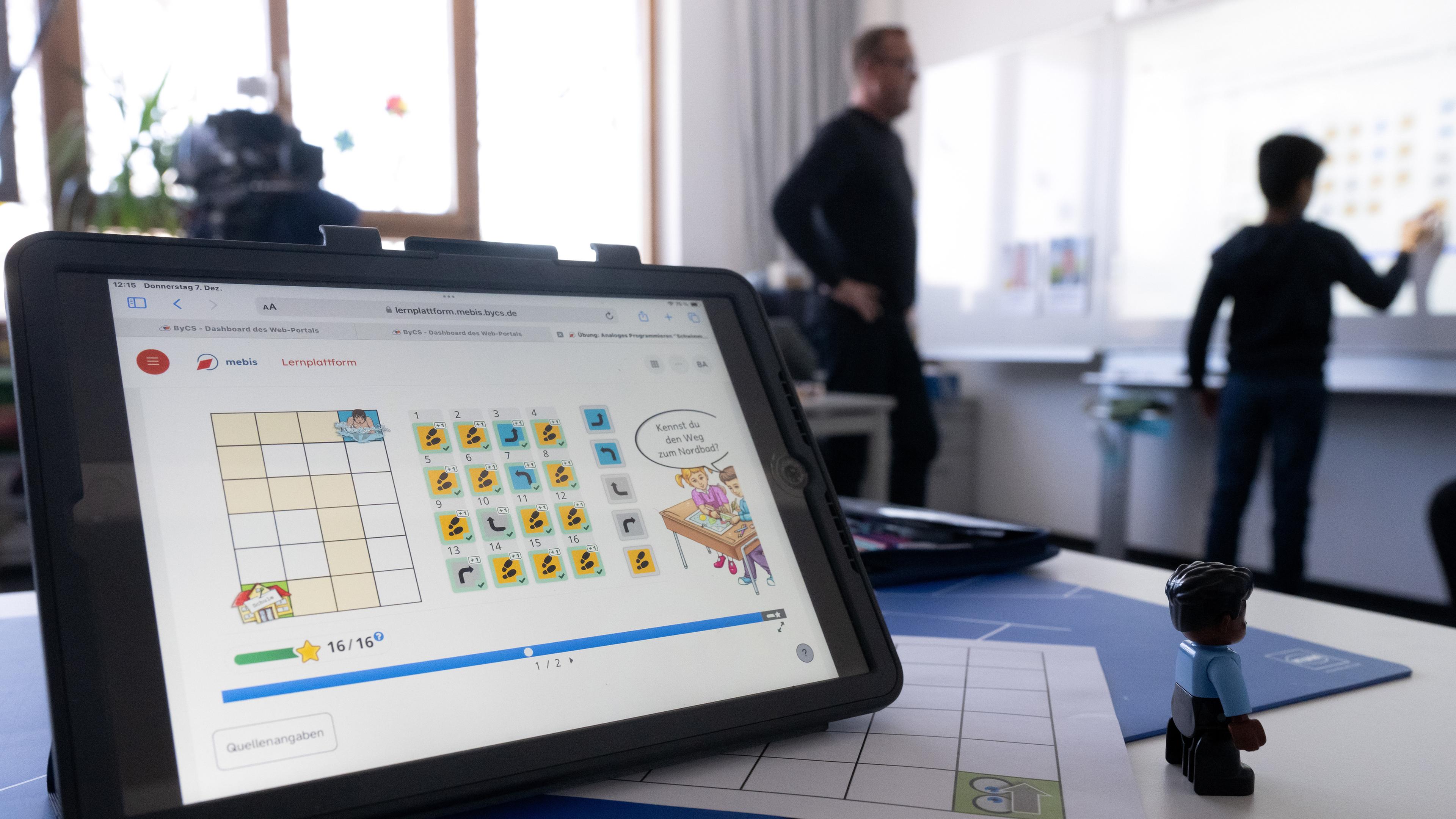 In einem Klassenzimmer stehen ein Schüler und eine Lehrkraft im Hintergrund an der Tafel, im Vordergrund steht ein Tablet auf dem Tisch.