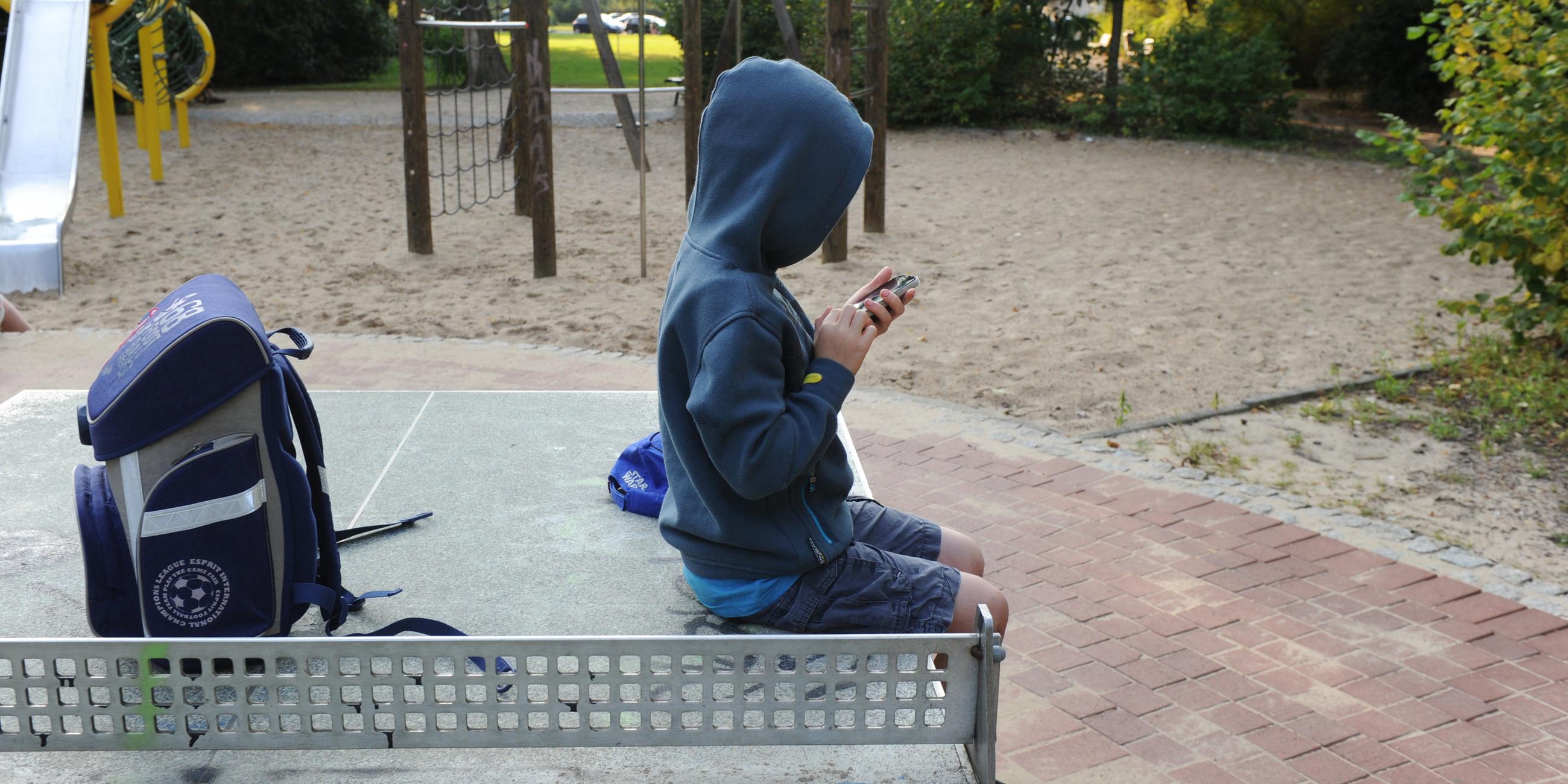 Ein Kind sitzt auf einer Tischtennisplatte auf einem Spielplatz und spielt mit einem Smartphone.