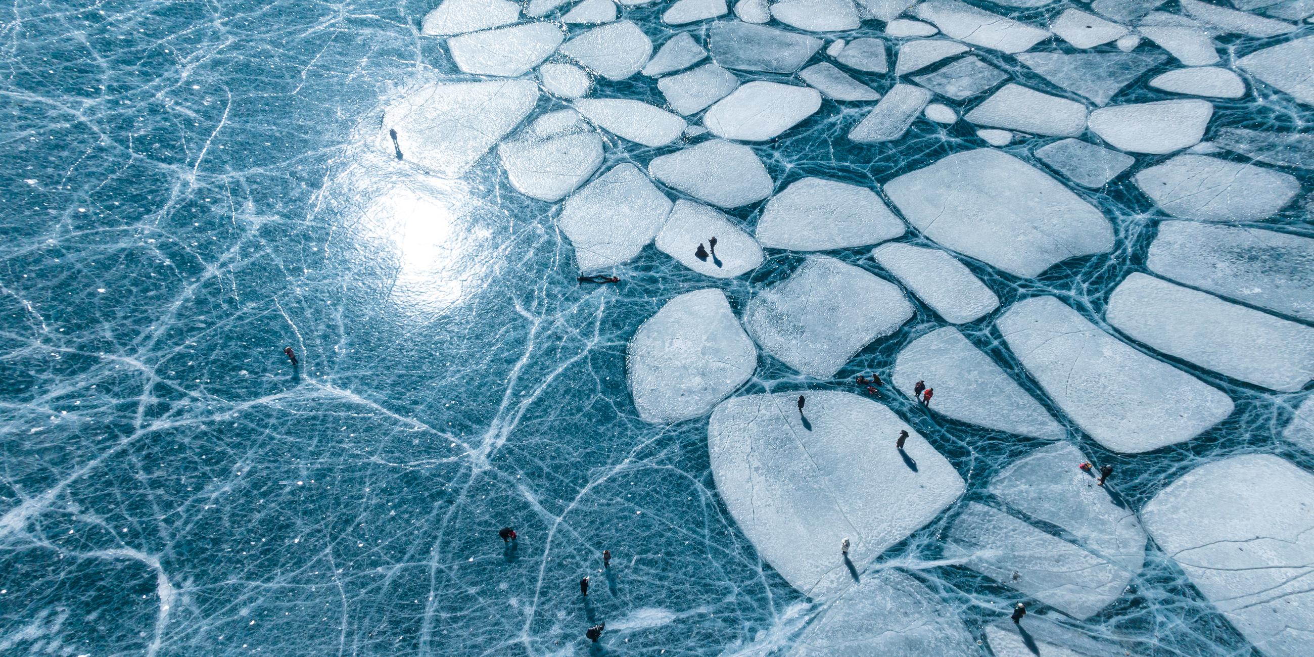 Ein Foto aus der Luft mit Blick auf einen großen See, der komplett mit Eis bedeckt ist. Auf der linken Seite ist das Eis einheitlich und das blaue Wasser scheint durch. Auf der rechten Seite besteht die Eisschicht aus dicken Mosaik-ähnlichen Eisschollen.