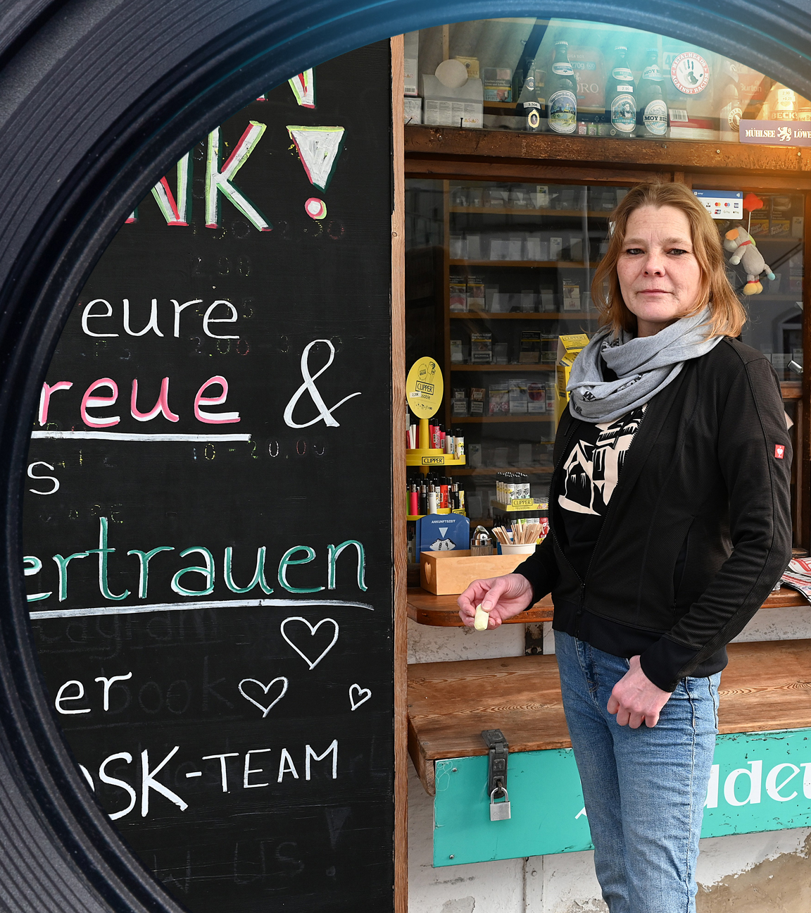 Daniela E. steht vor ihrem Kiosk und schaut in die Kamera. Neben ihr ist eine Tafel zu sehen, auf der Abschiedsworte an ihre Kundschaft stehen.