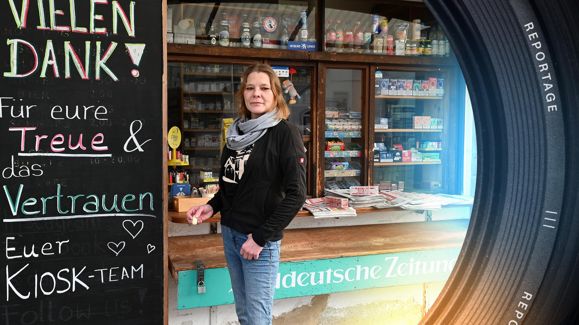 Daniela E. steht vor ihrem Kiosk und schaut in die Kamera. Neben ihr ist eine Tafel zu sehen, auf der Abschiedsworte an ihre Kundschaft stehen.