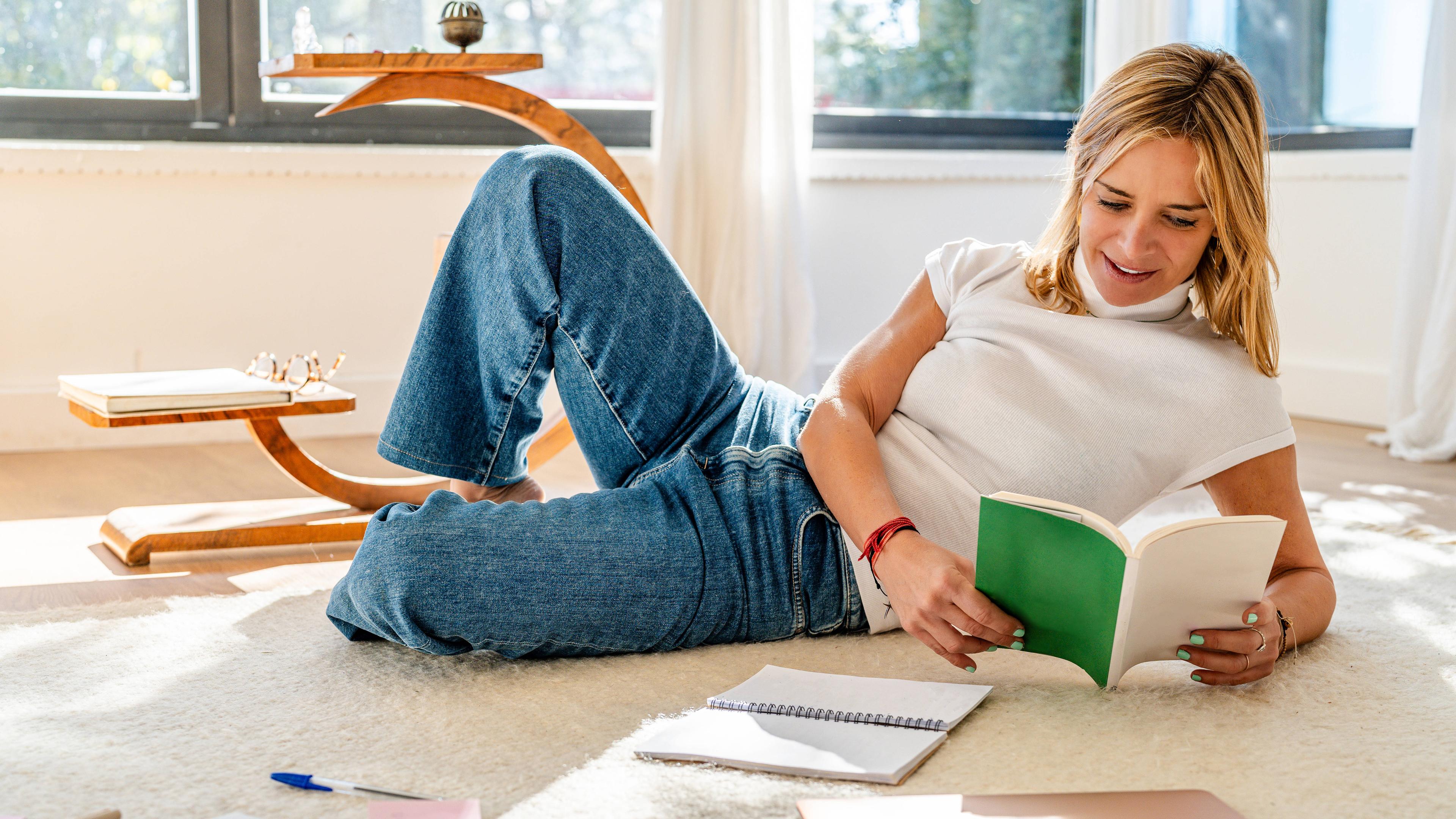 Frau liegt auf dem Boden und hält ein Buch in der Hand, um sie herum liegen Arbeitsmaterialen.