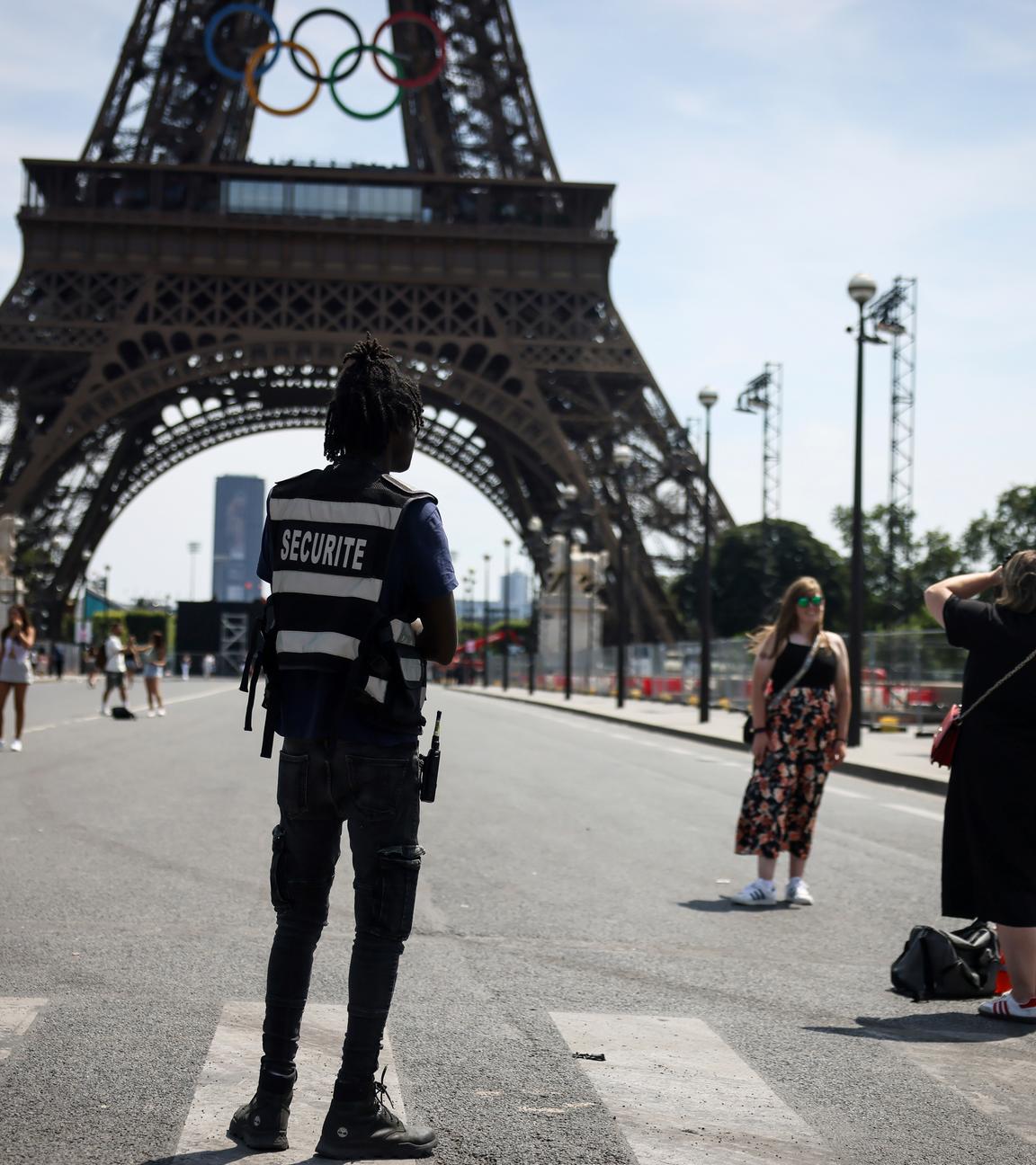 20.07.2024, Frankreich, Paris: Ein Sicherheitsbeamter beobachtet Menschen, die vor dem Eiffelturm fotografiert werden.