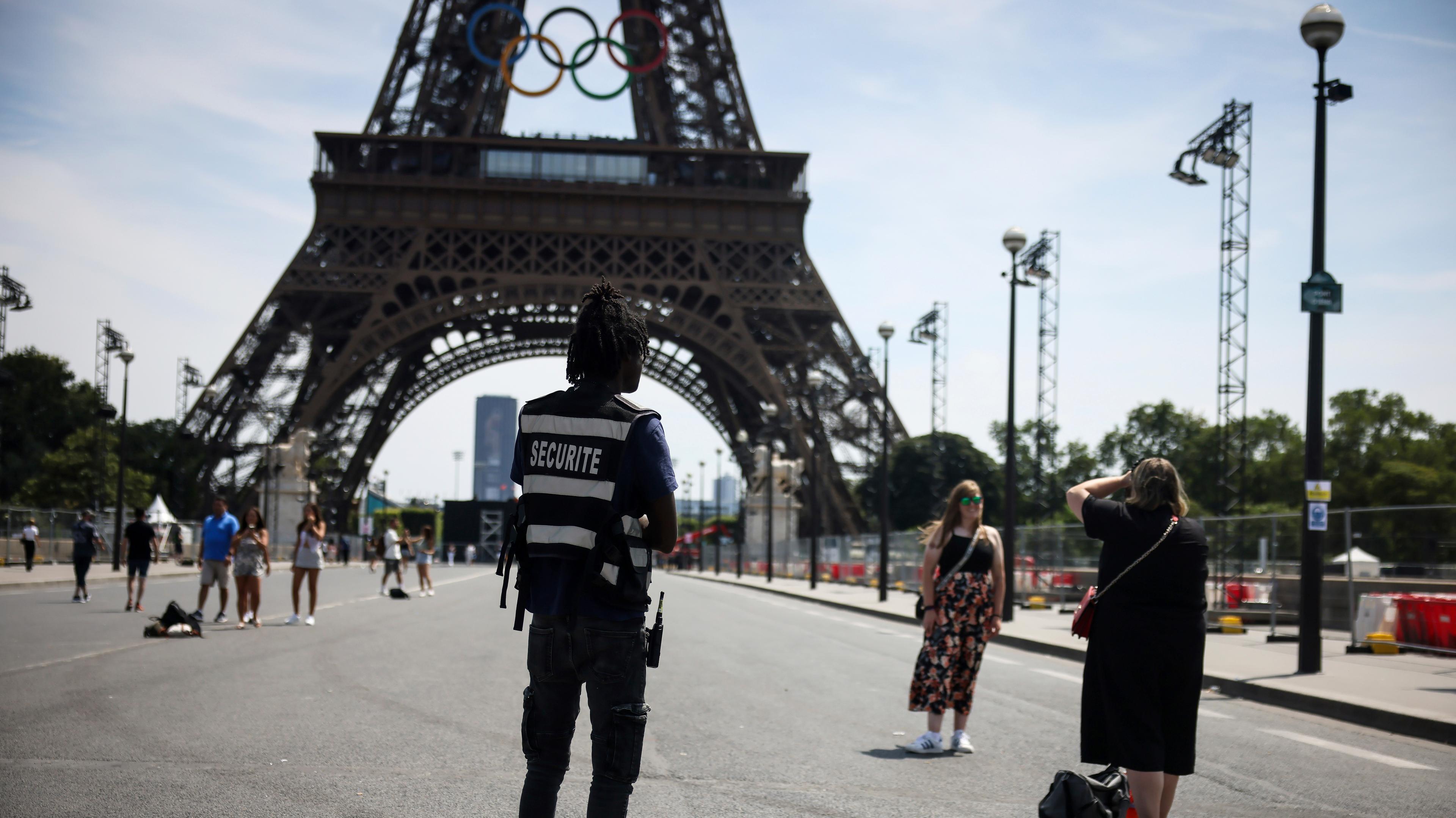20.07.2024, Frankreich, Paris: Ein Sicherheitsbeamter beobachtet Menschen, die vor dem Eiffelturm fotografiert werden.