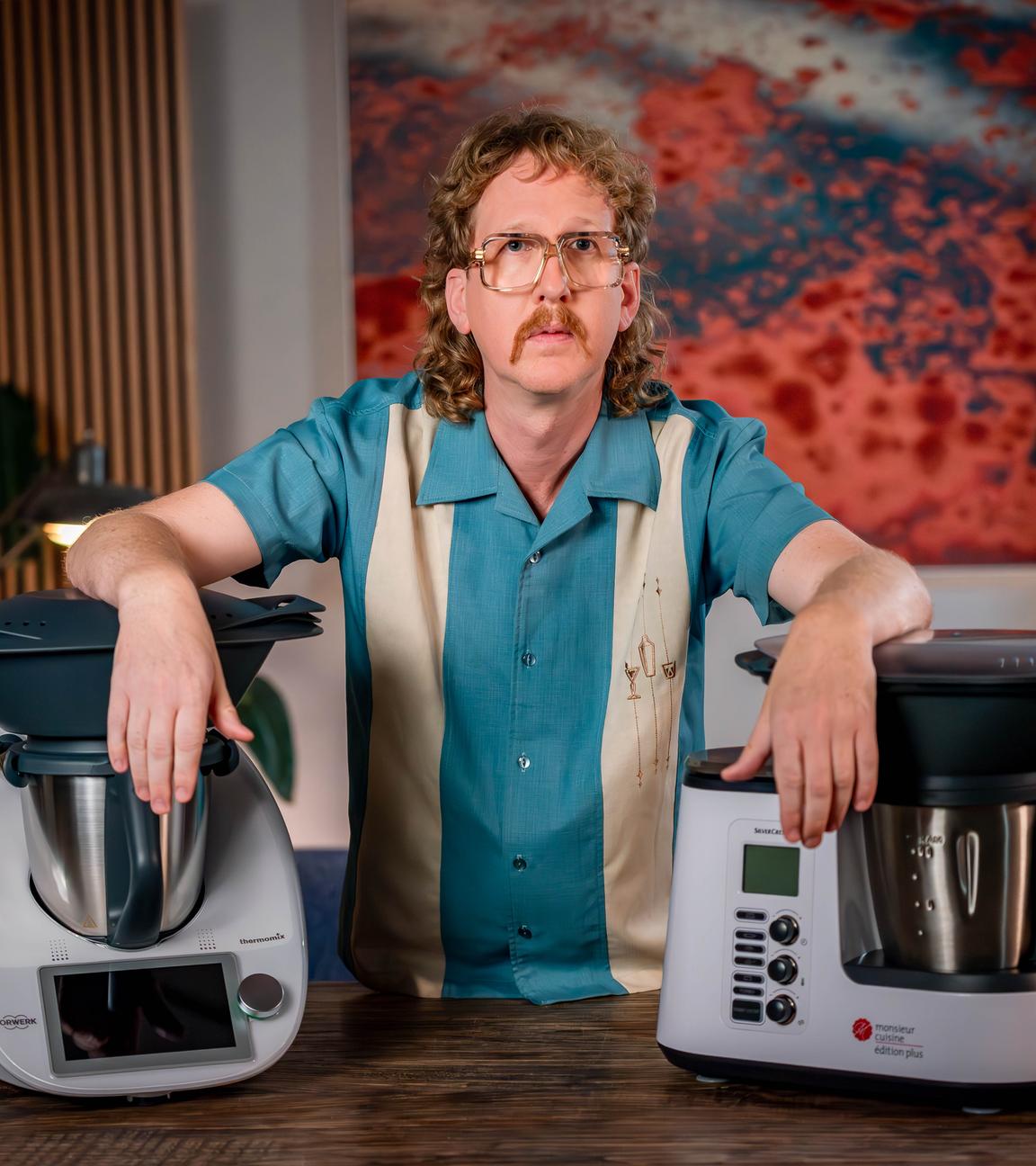 Uke Bosse schaut ernst in die Kamera, seine Arme stützen sich links auf einen Thermomix und rechts auf eine "Monsieur Cuisine"-Maschine.