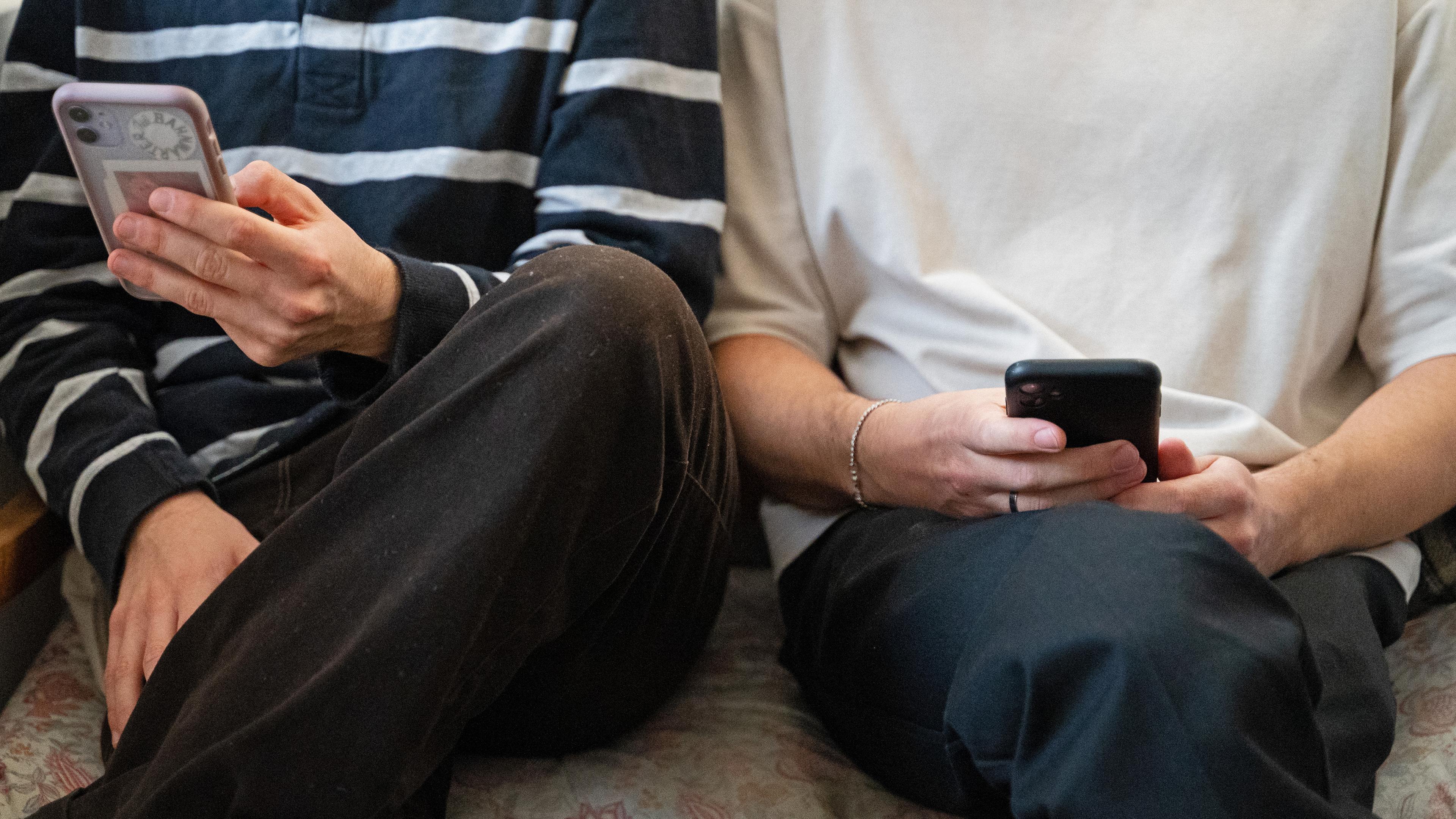 Baden-Württemberg, Stuttgart: Zwei junge Männer sitzen nebeneinander auf einer Bank und halten ihr Smartphones in den Händen.