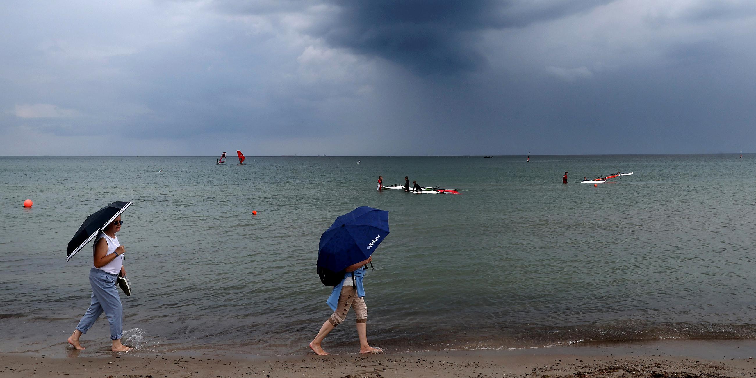 Über der Ostsee ziehen dunkle Regenwolken auf, während zwei Frauen mit Regenschirmen am Strand unterwegs sind