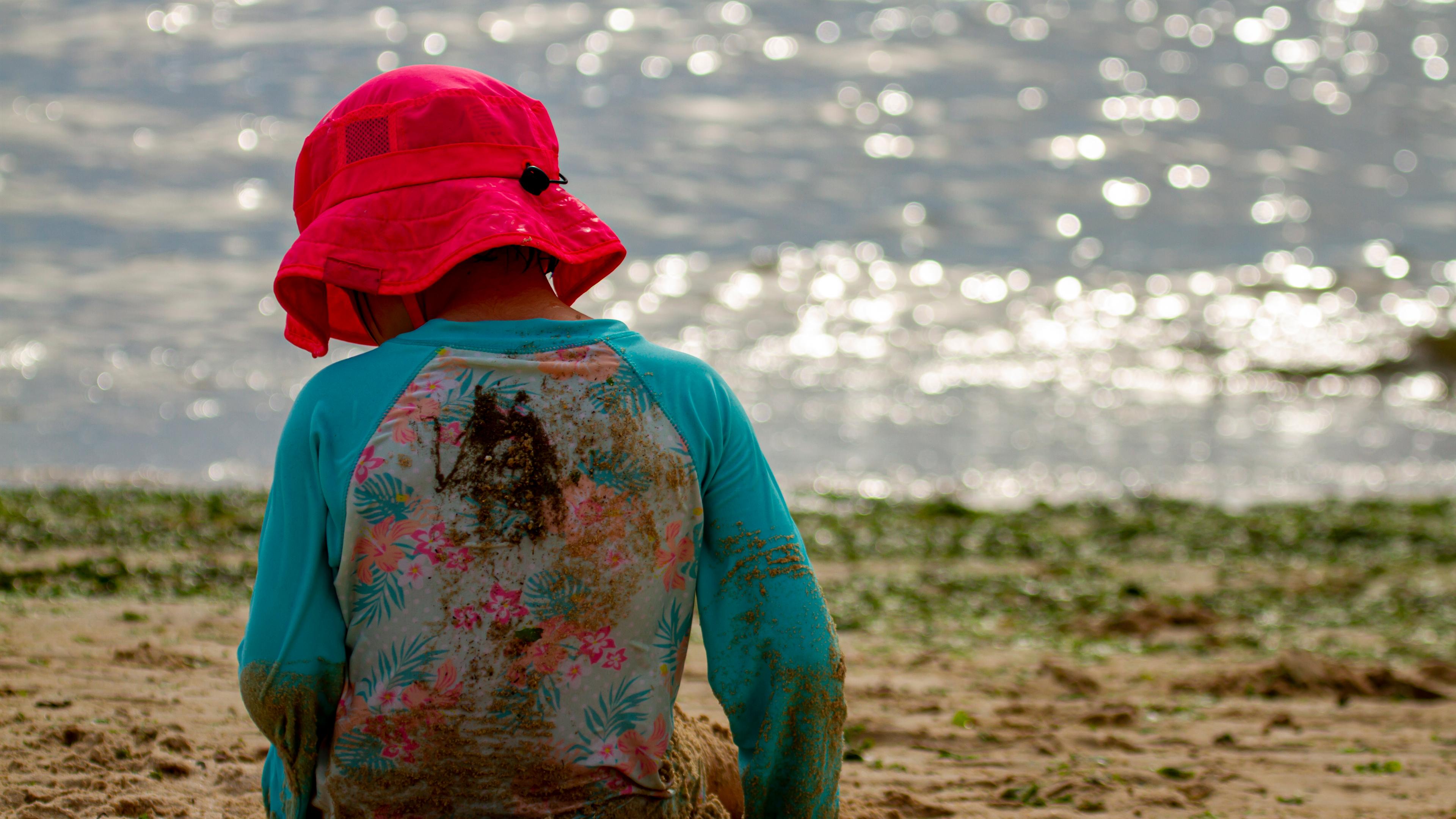 Ein junges Mädchen mit Sonnenhut und langärmeliger Kleidung sitzt am Seeufer im Sand. 