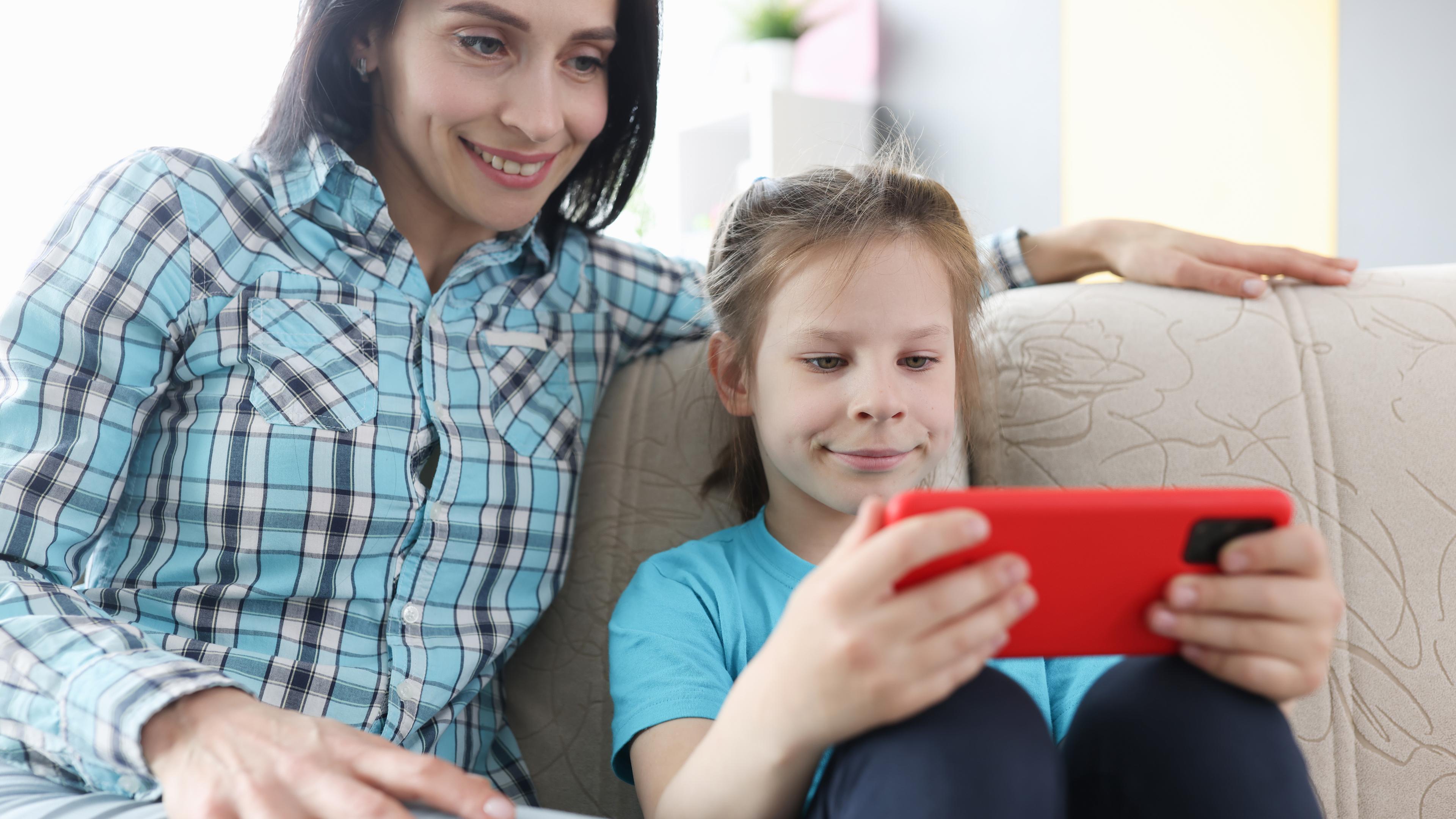 Eine erwachsene Frau mit braunen Haaren und ein junges Mädchen mit blonden Haaren sitzen auf einer Couch und schauen zusammen auf ein orangenes Smartphone.