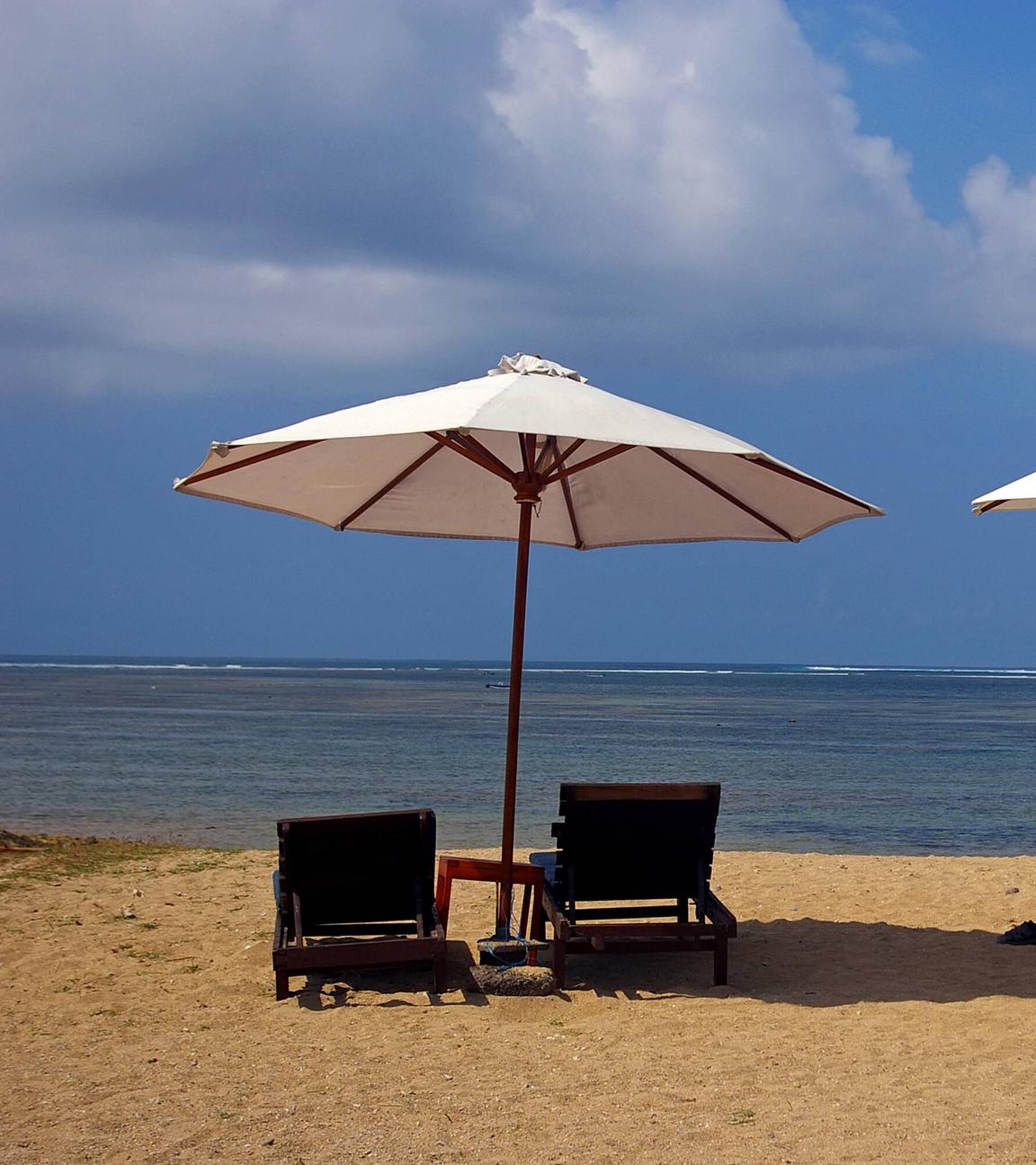 Zwei leere Strandliegen mit Sonnenschirmen stehen an einem Strand mit Blick auf das Meer.