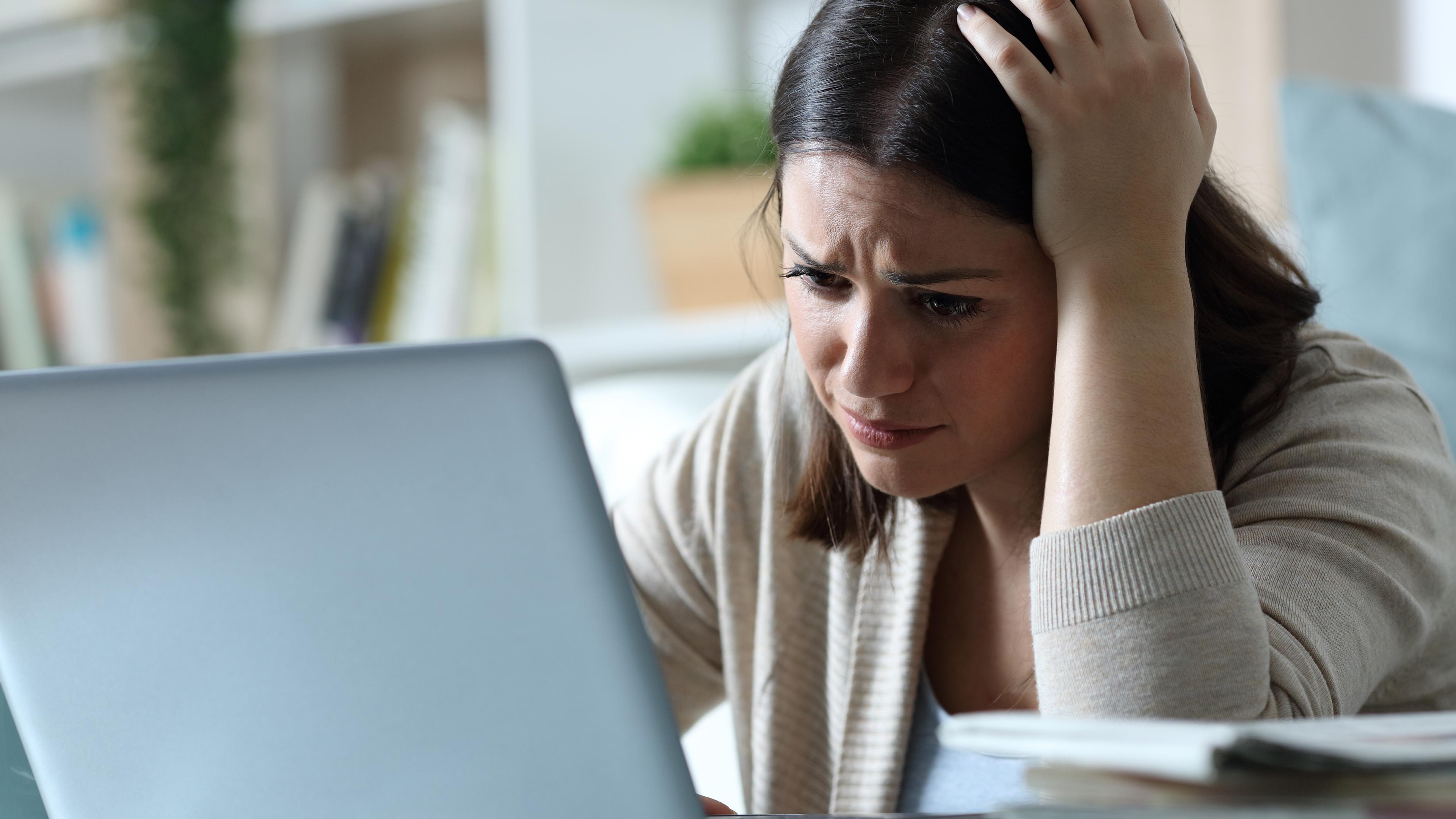 Eine junge Frau stützt ihren Kopf auf ihrer Hand auf und schaut verzweifelt auf einen Laptop-Bildschirm.