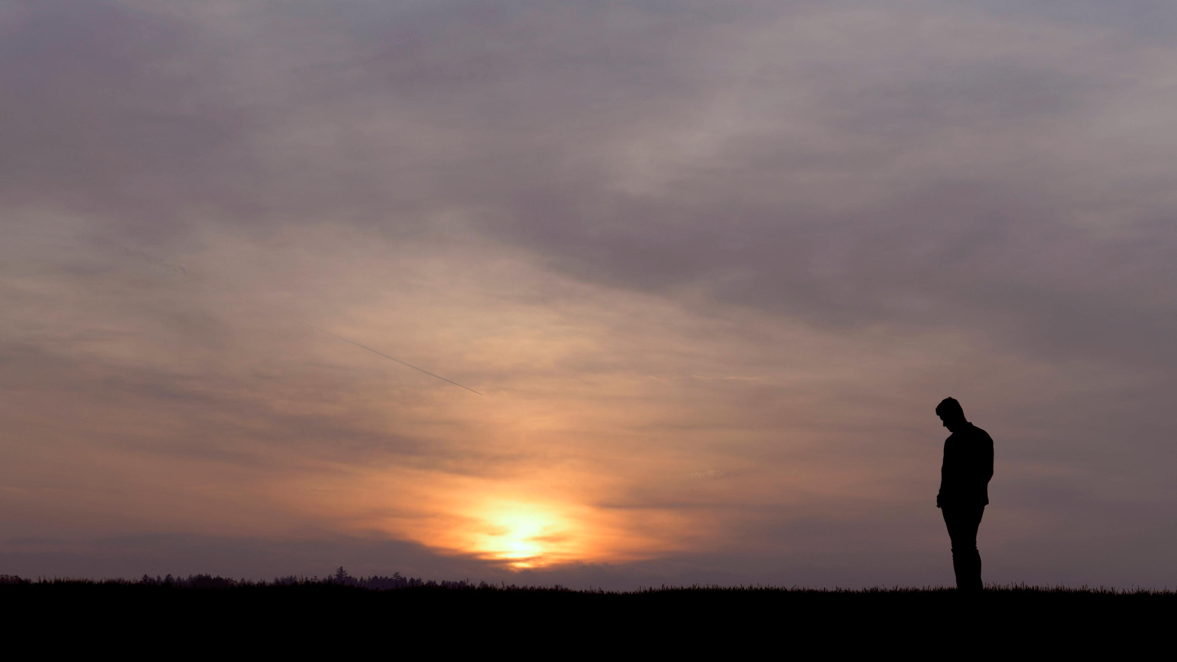 Junger Mann steht in der Abenddämmerung auf einem Feld, am Horizont ist der Sonnenuntergang zu sehen.