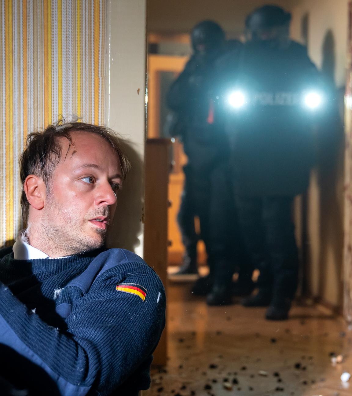 Ein verschwitzter Mann mit einer Waffe in der Hand versteckt sich hinter einer Wand, während im angrenzenden Flur Spezialeinsatzkräfte vorrücken.