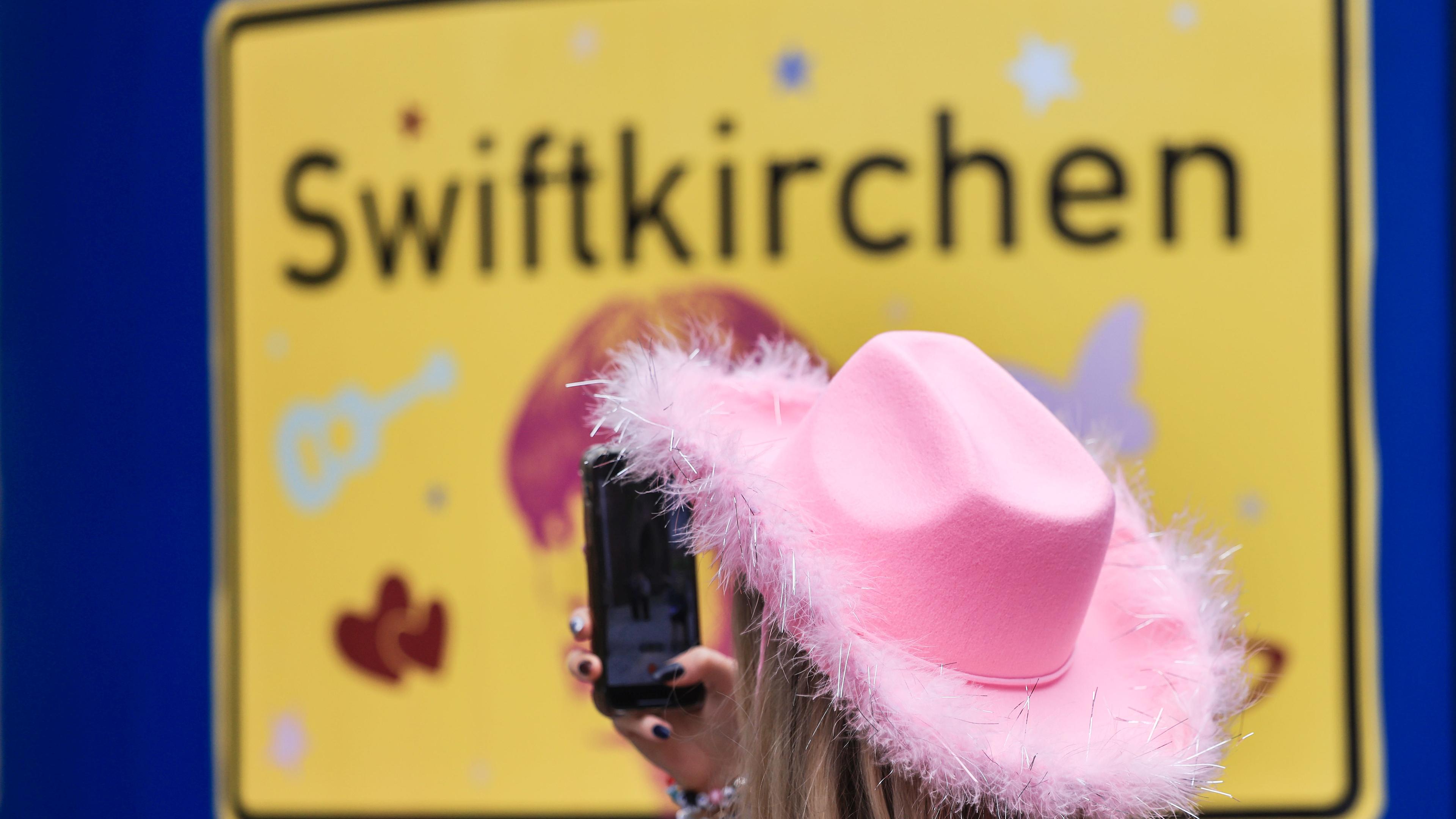 "Swiftie" vor Stadtschild mit der Aufschrift "Swiftkirchen"