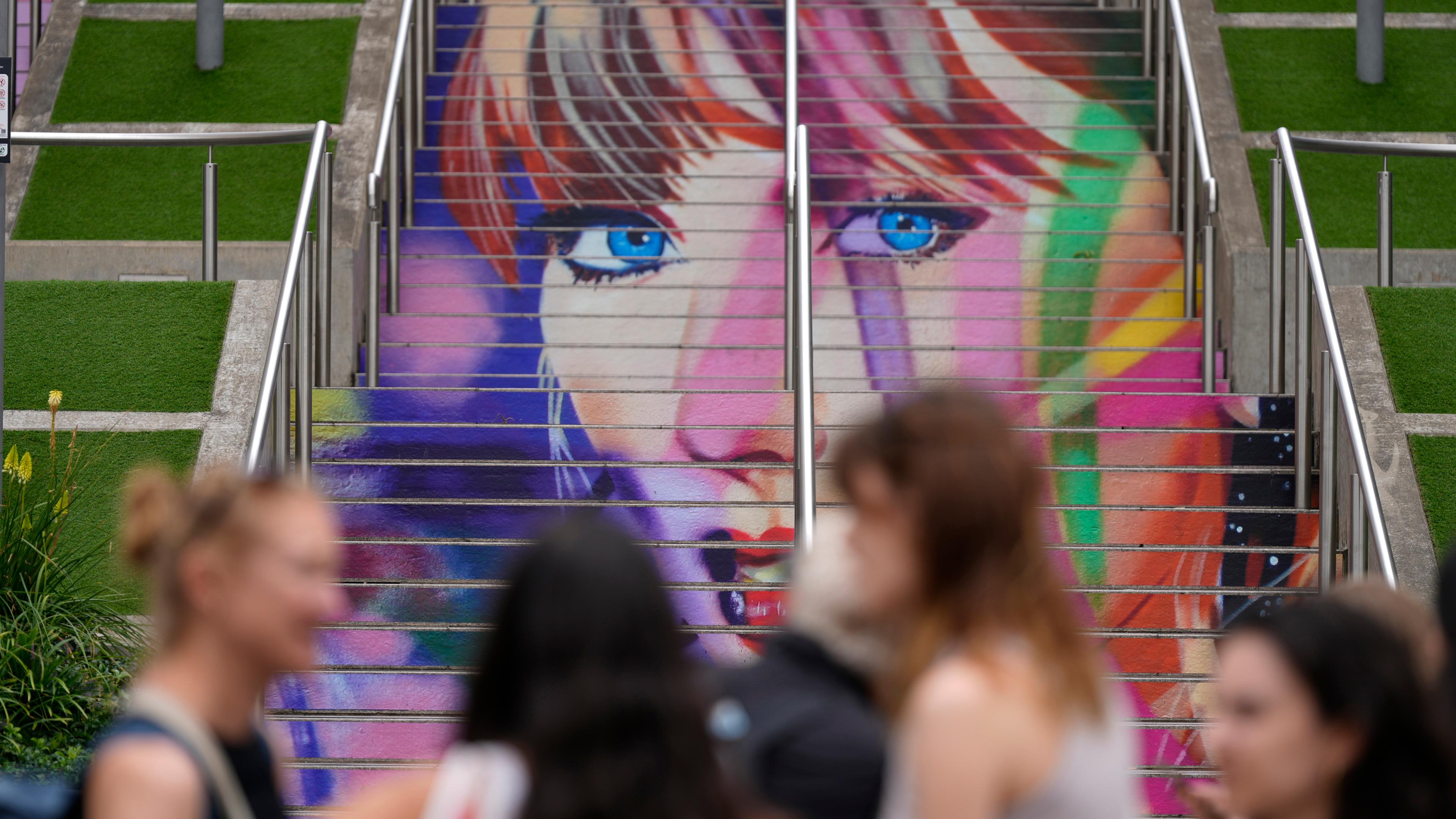 Fans posieren vor einem Porträt von Taylor Swift, das auf einer Treppe im Wembley-Stadion in London gemalt ist. 