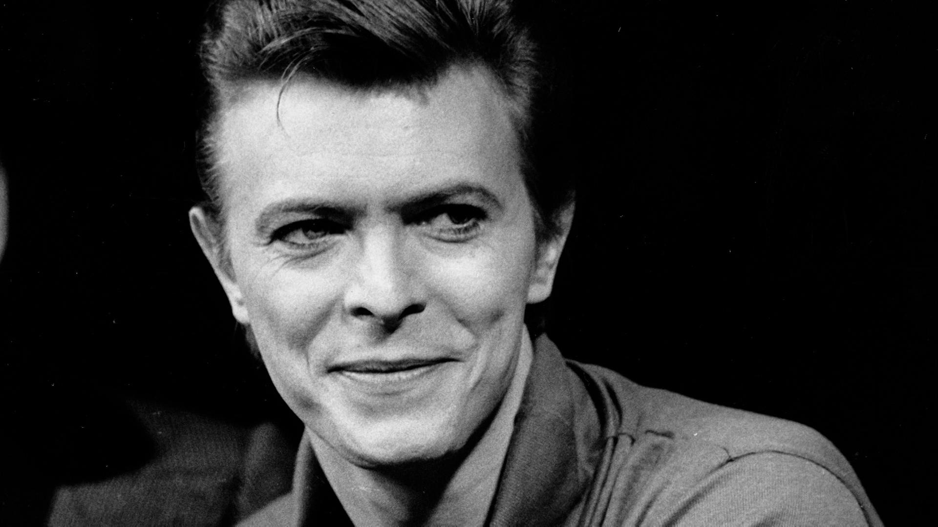  Schwarz-Weiß-Portrait von David Bowie in einer Interview-Situation. Er blickt rechts an der Kamera vorbei.