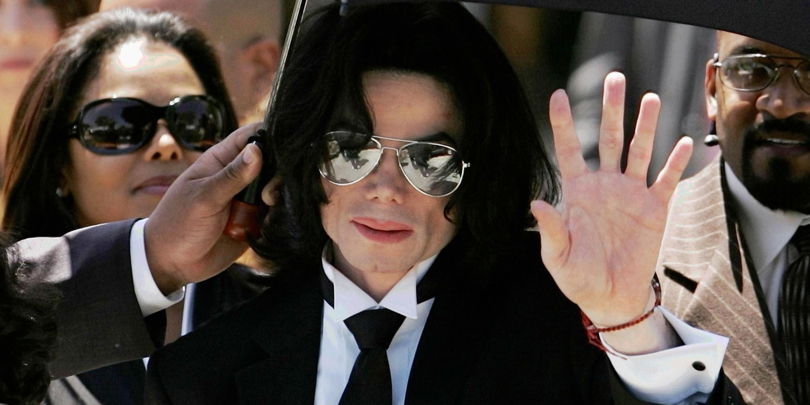 Michael Jackson mit Anzug und Krawatte inmitten einer Menschengruppe. Eine Hand hält ihm von links einen Sonnenschirm über den Kopf. Jackson hat die linke Hand erhoben.