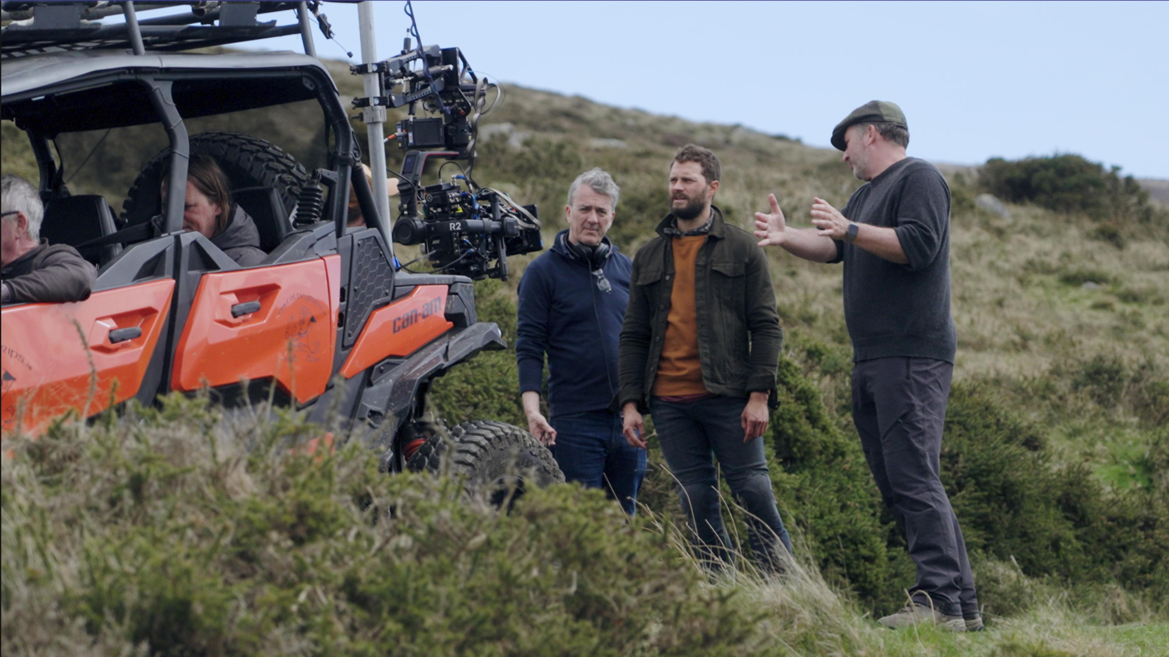 "The Tourist - Irisches Blut: Making-of, Teil 1": Jamie Dornan steht mit zwei Männern hinter einem roten Strandbuggy in einer hügeligen Dünenlandschaft. Auf dem Buggy ist eine Kamera montiert.