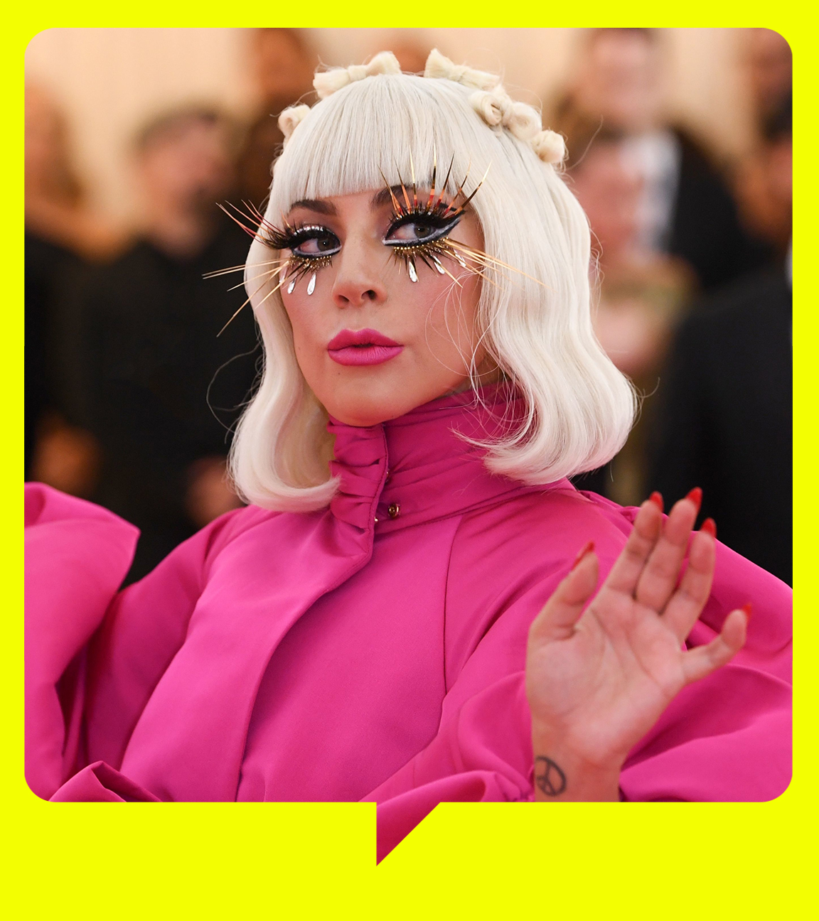  Lady Gaga trägt ein rosafarbenes Kleid mit Puffärmeln. Hellblonde Haare reichen ihr bis zur Schulter und sie trägt lange künstliche Wimpern. Im Hintergrund sind Fotografen und ein Mann im Smoking zu sehen.