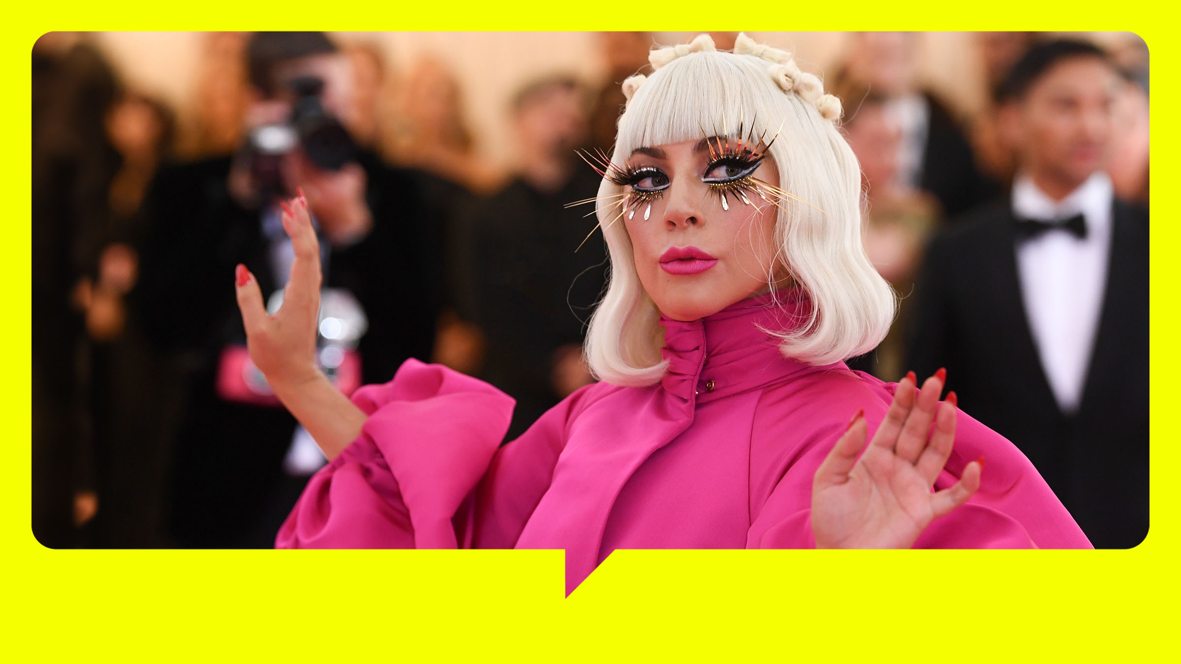  Lady Gaga trägt ein rosafarbenes Kleid mit Puffärmeln. Hellblonde Haare reichen ihr bis zur Schulter und sie trägt lange künstliche Wimpern. Im Hintergrund sind Fotografen und ein Mann im Smoking zu sehen.