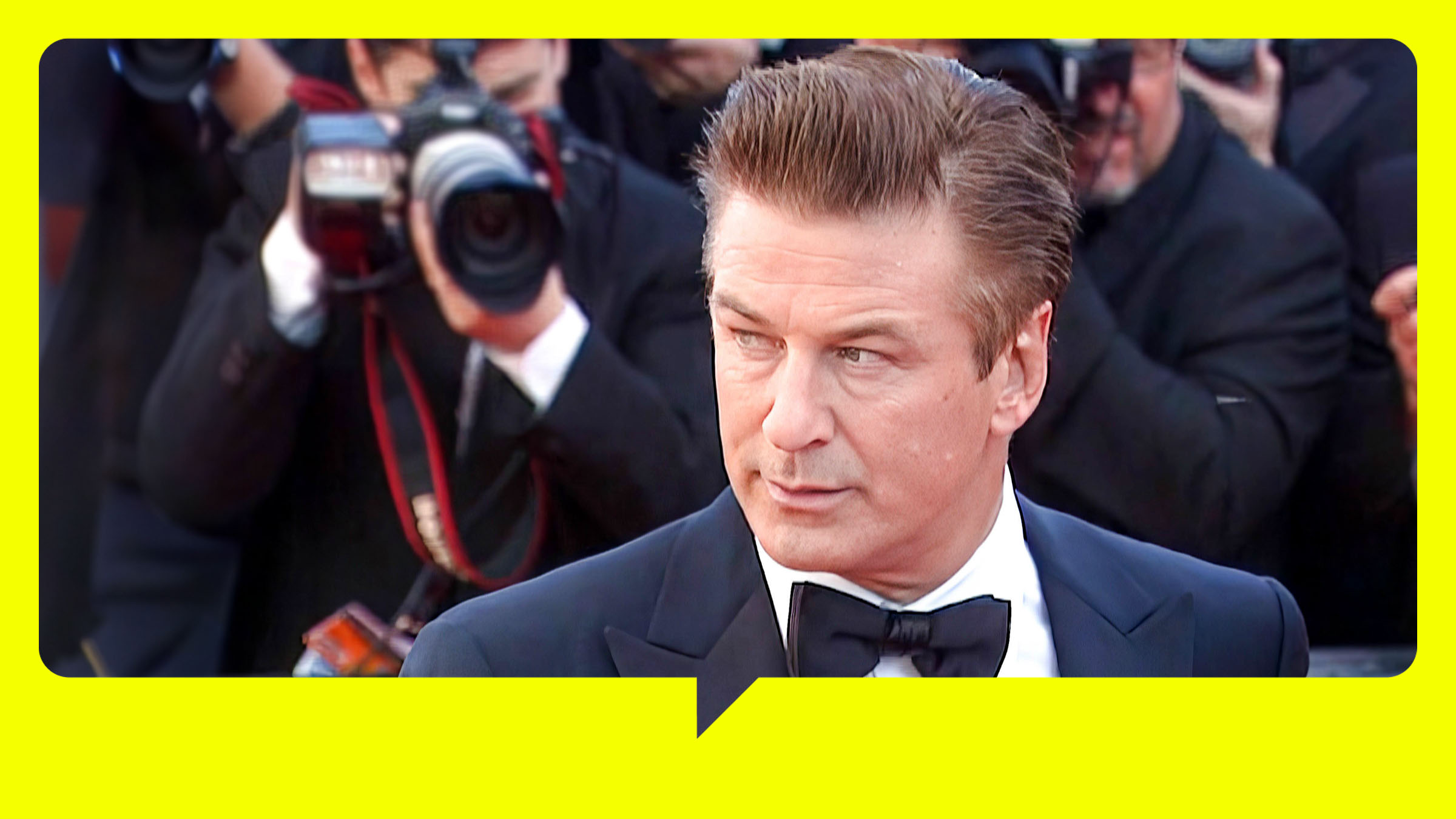 Alex Baldwin steht in Anzug und Fliege neben seiner dunkelhaarigen Frau. Beide schauen zur Seite, hinter ihnen stehen Fotografen.