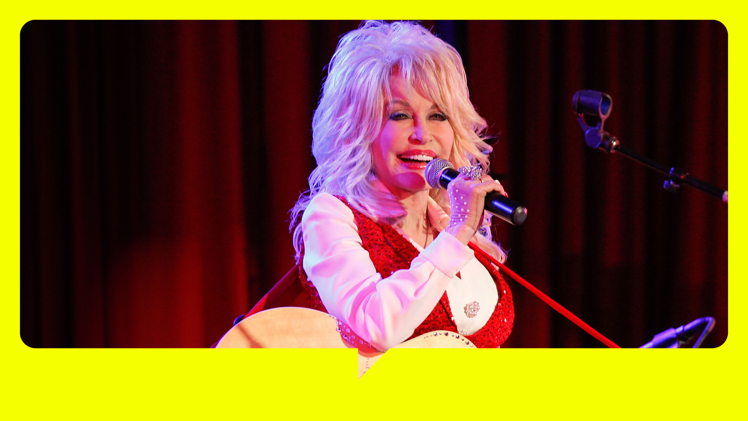 Dolly Parton hält in der linken Hand eine Gitarre. Sie steht vor einem roten Vorhang und singt in ein Mikrofon.