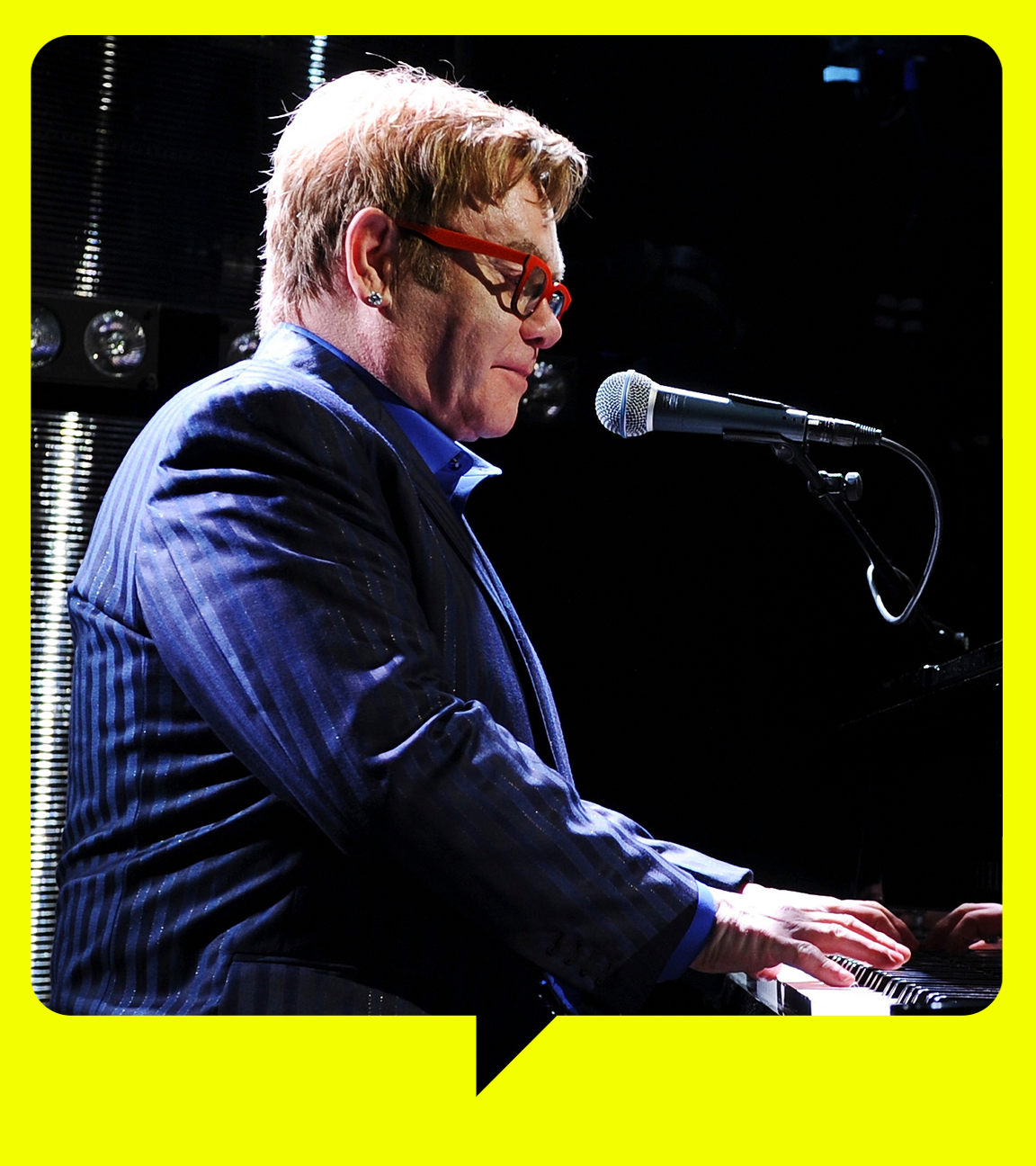 Elton John trägt eine rote Sonnenbrille und ein gestreiftes blaues Sakko. Er spielt Klavier.