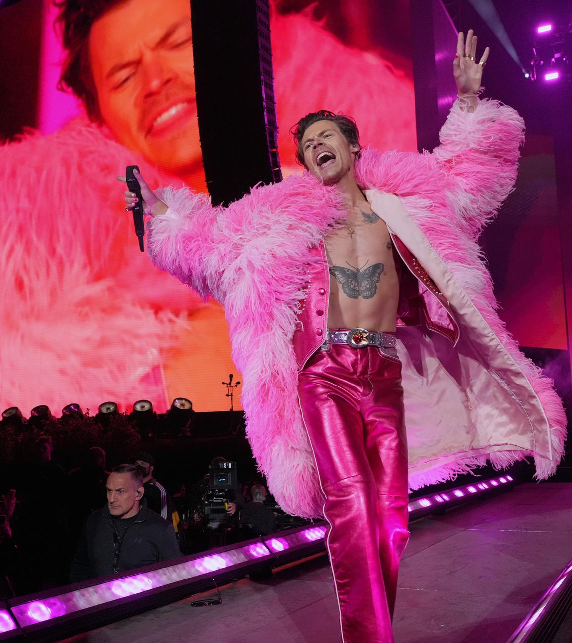 Harry Styles in pinker Metallic-Hose und pinkem Plüschmantel auf einer Bühne. Im Hintergrund Scheinwerfer und Übertragungsleinwand.