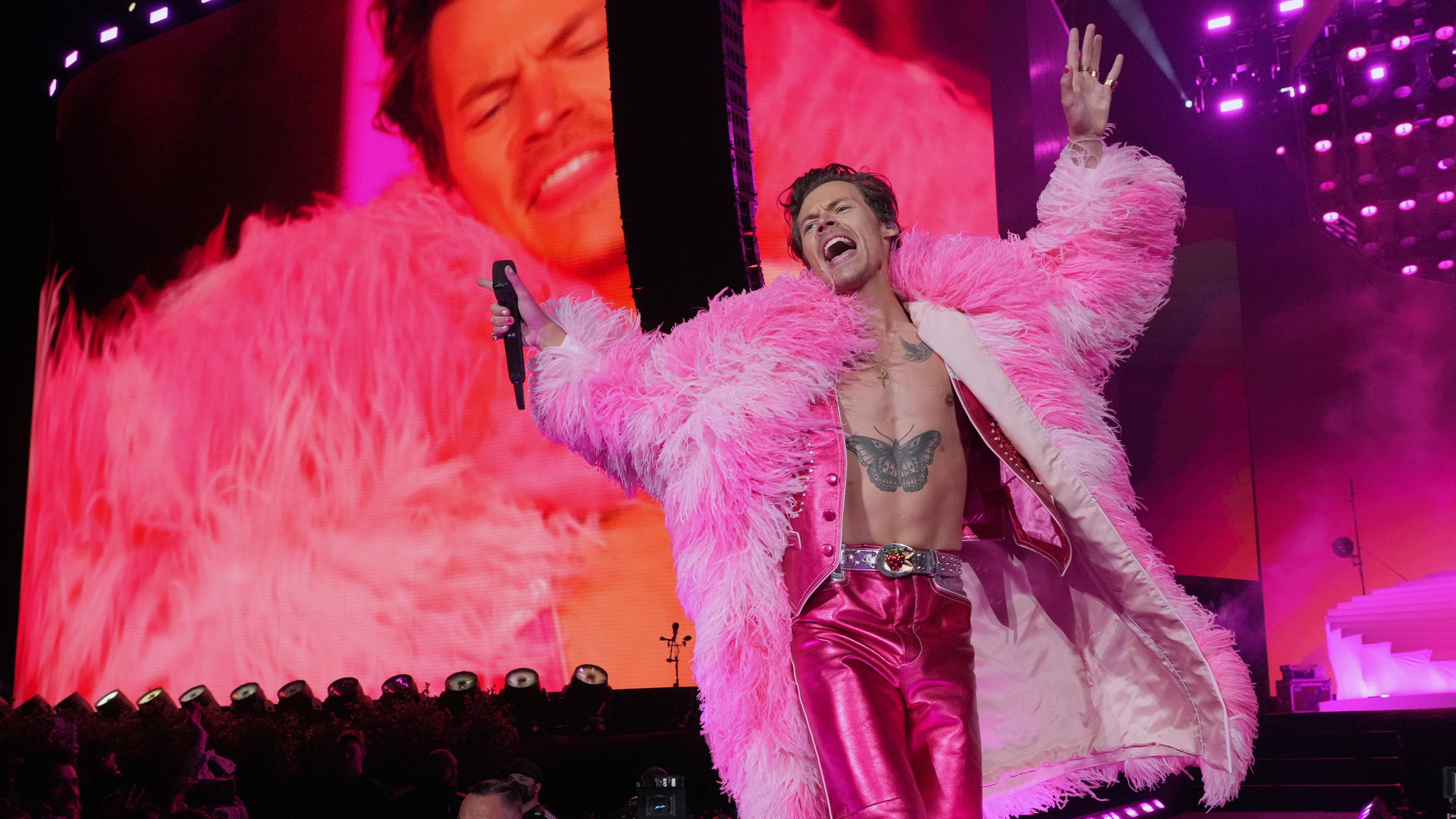 Harry Styles in pinker Metallic-Hose und pinkem Plüschmantel auf einer Bühne. Im Hintergrund Scheinwerfer und Übertragungsleinwand.