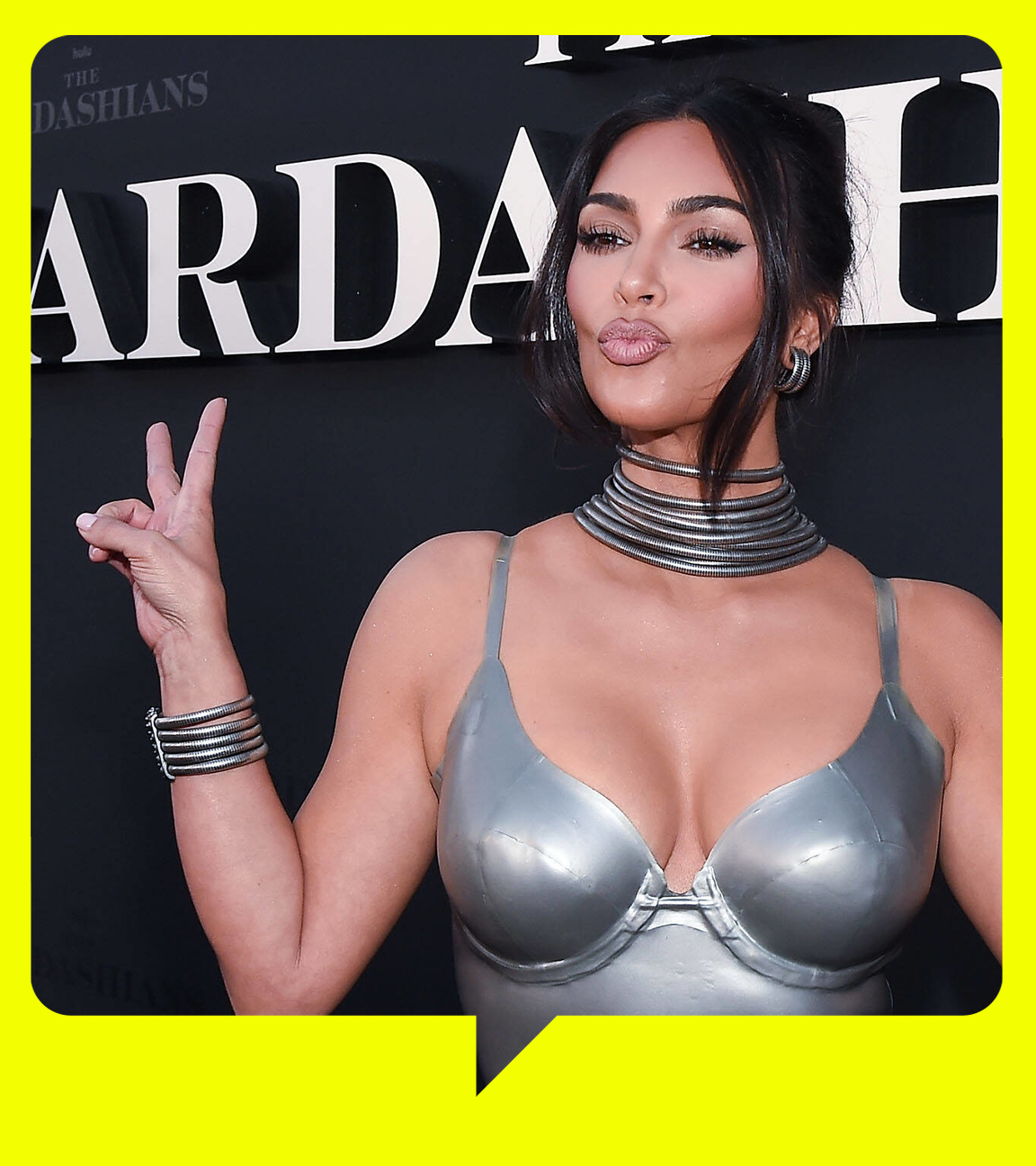 Kim Kardashian steht vor einer schwarzen Wand mit der Aufschrift "The Kardashians". Sie trägt ein silberfarbenes Kleid und silbernen Schmuck. Sie macht einen Kussmund und zeigt mit beiden Händen das Peace-Zeichen.