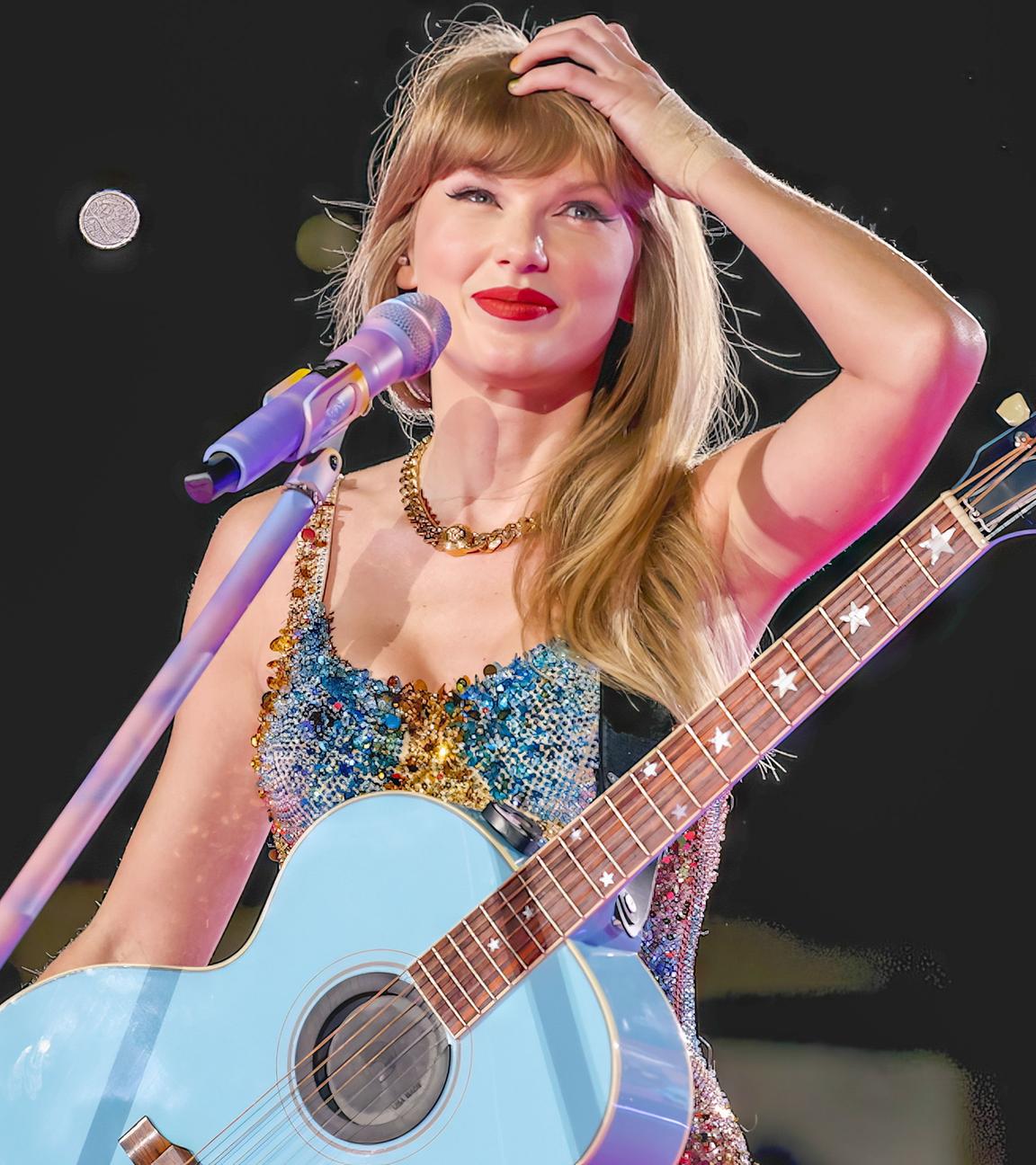 Taylor Swift steht mit Gitarre an einem Mikrofon. Sie lächelt und fasst sich an den Kopf.