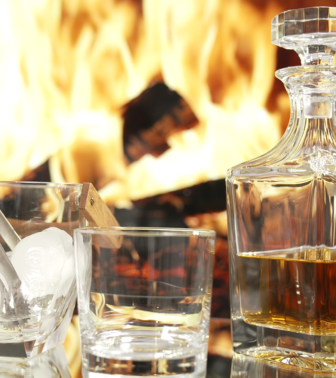 Whiskey in Karaffe mit zwei Gläsern. In einem Glas sind Eiswürfel. Im Hintergrund lodert ein Kaminfeuer.