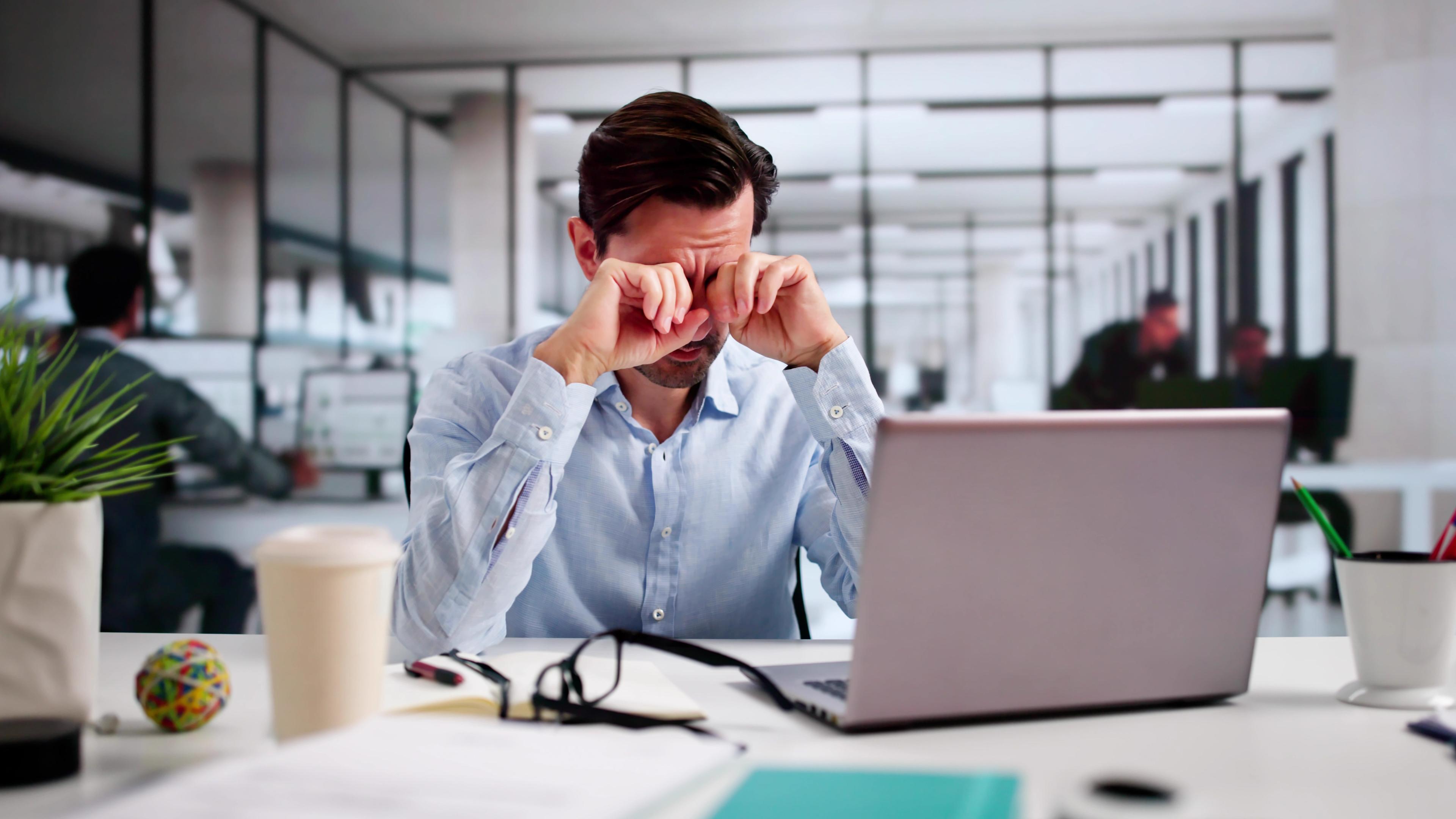 Ein Mann, der im Büro an einem Laptop arbeitet, reibt sich die Augen. 