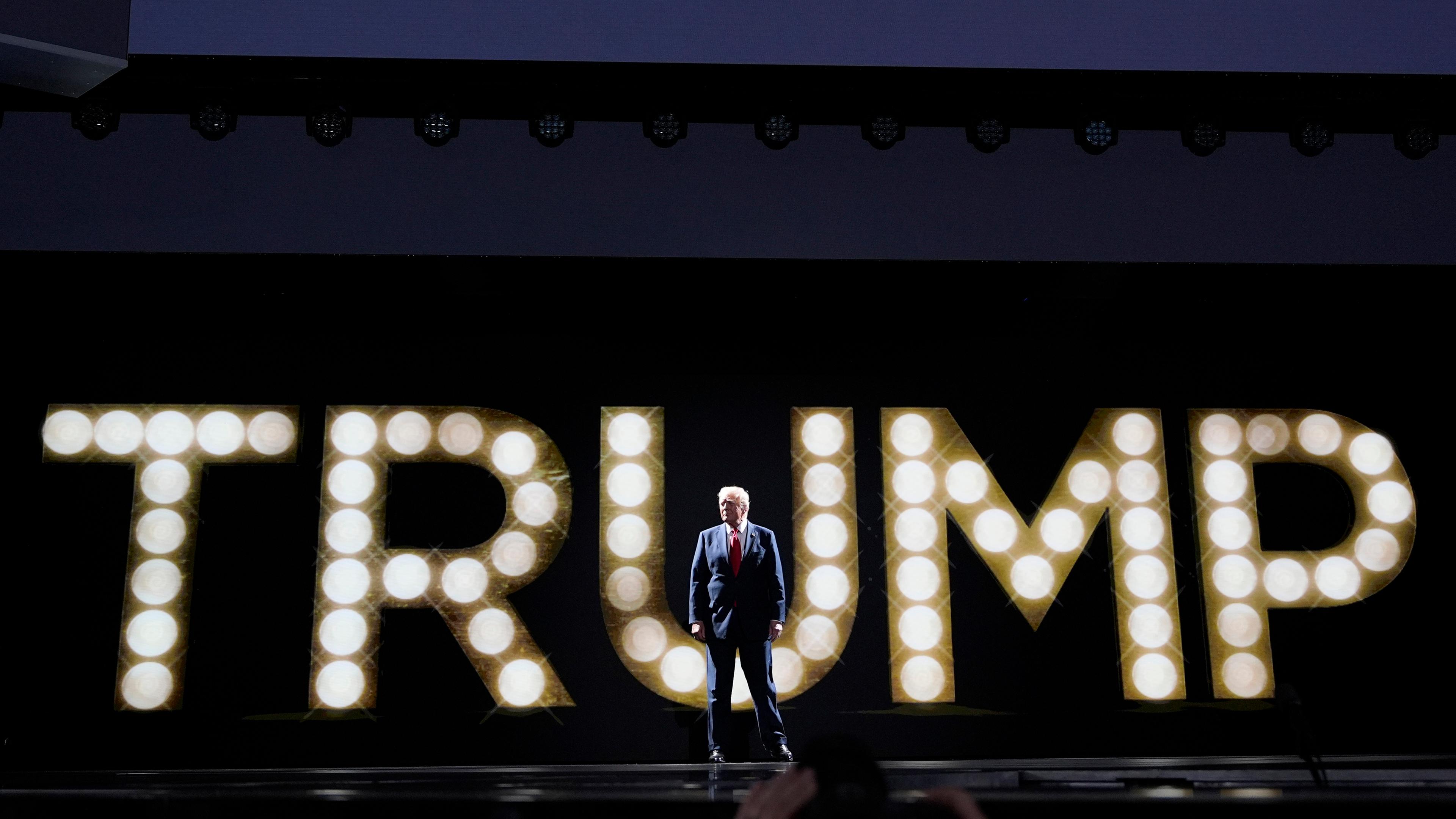 In riesigen Leuchtbuchstaben ist Trump zu lesen, davor steht im Anzug Donald Trump 