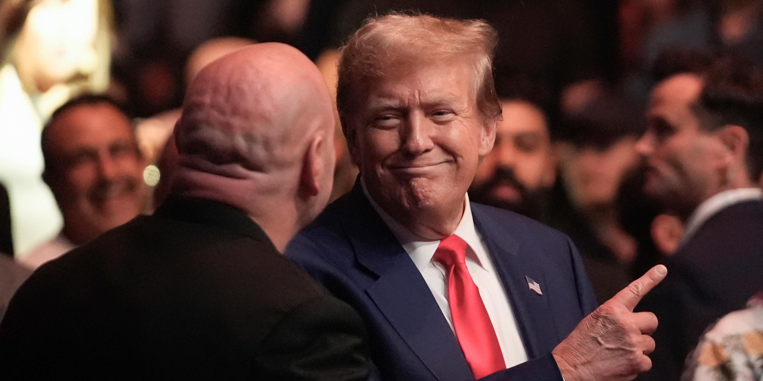 Donald Trump steht im blauen Anzug mit roter Krawatte in einer Menge und zeigt mit dem Zeigefinger nach oben.