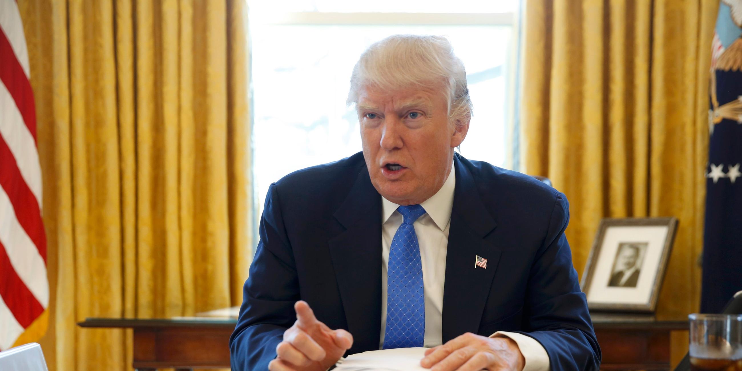 Donald Trump sitzt im Oval Office hinter dem großen Schreibtisch und zeigt mit erhobenem Zeigefinger in Richtung Kamera.