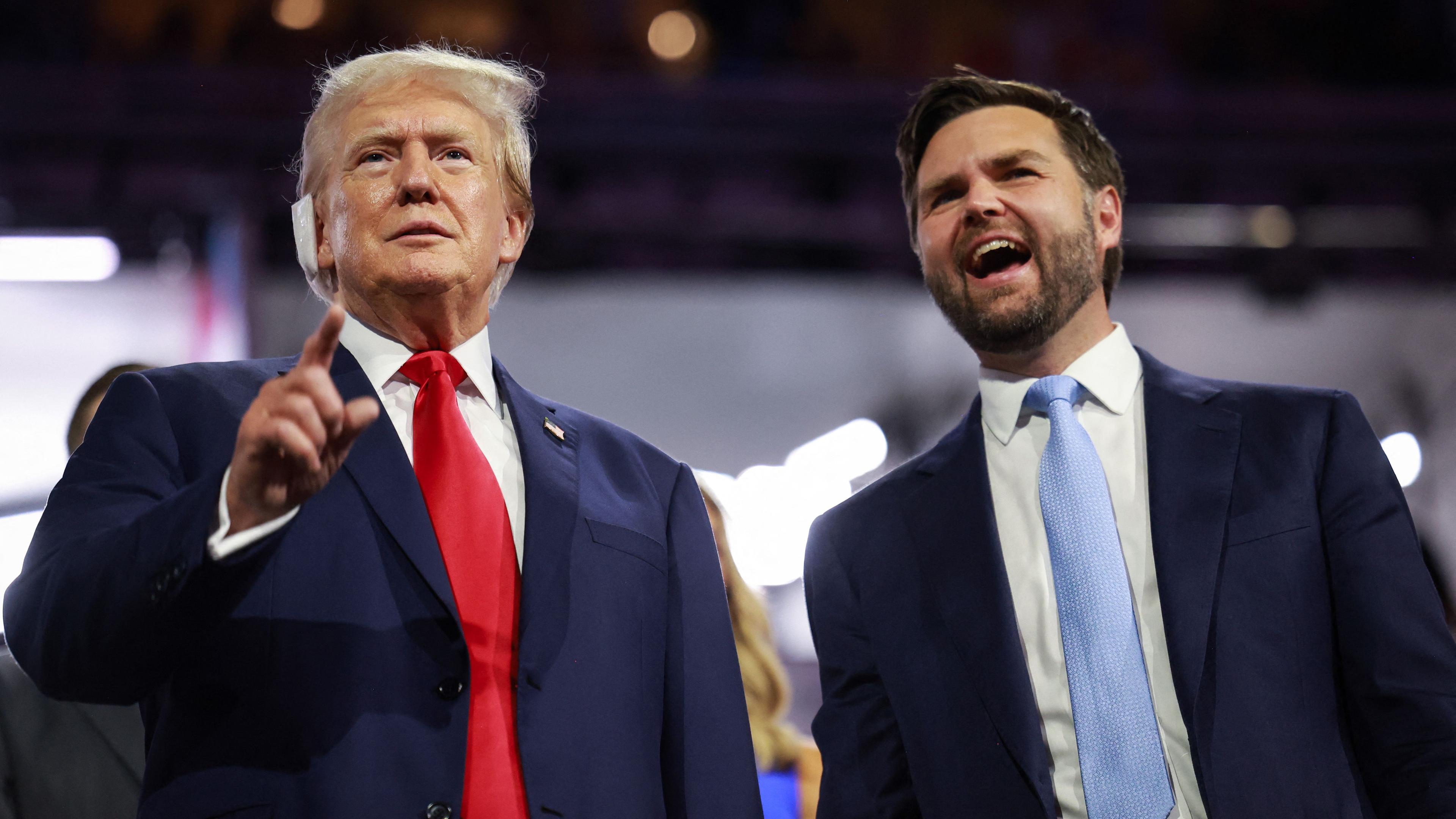 Donald Trump und J.D. Vance stehen im Anzug auf einer Tribüne. Trump zeigt in die Menge und Vance lächelt neben ihm. 