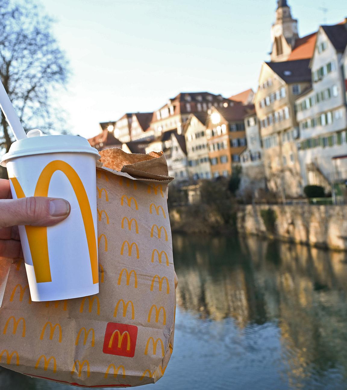 Eine McDonald's To-Go-Tüte und ein To-Go-Becher werden auf der Neckarbrücke in Tübingen vor der Kulisse der Altstadt gehalten (gestellte Szene)