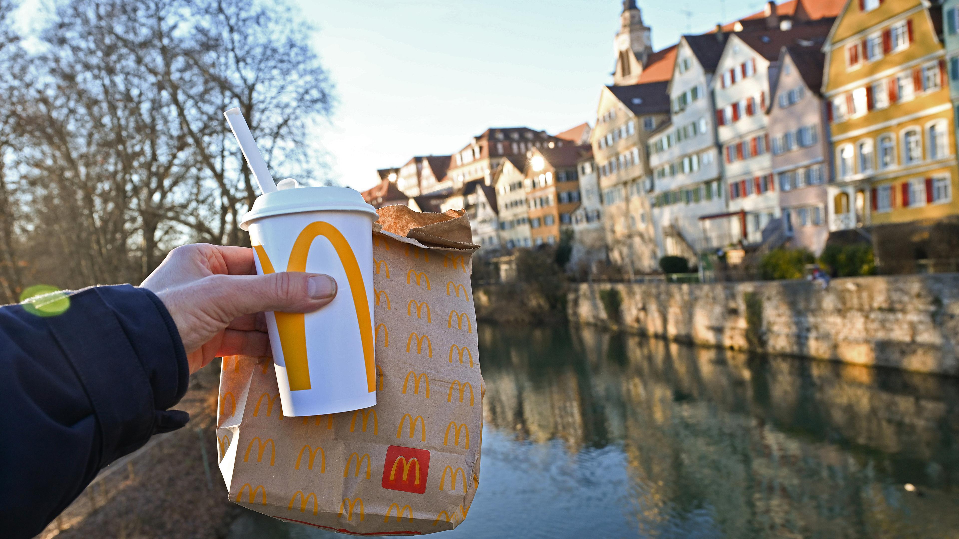 Eine McDonald's To-Go-Tüte und ein To-Go-Becher werden auf der Neckarbrücke in Tübingen vor der Kulisse der Altstadt gehalten (gestellte Szene)