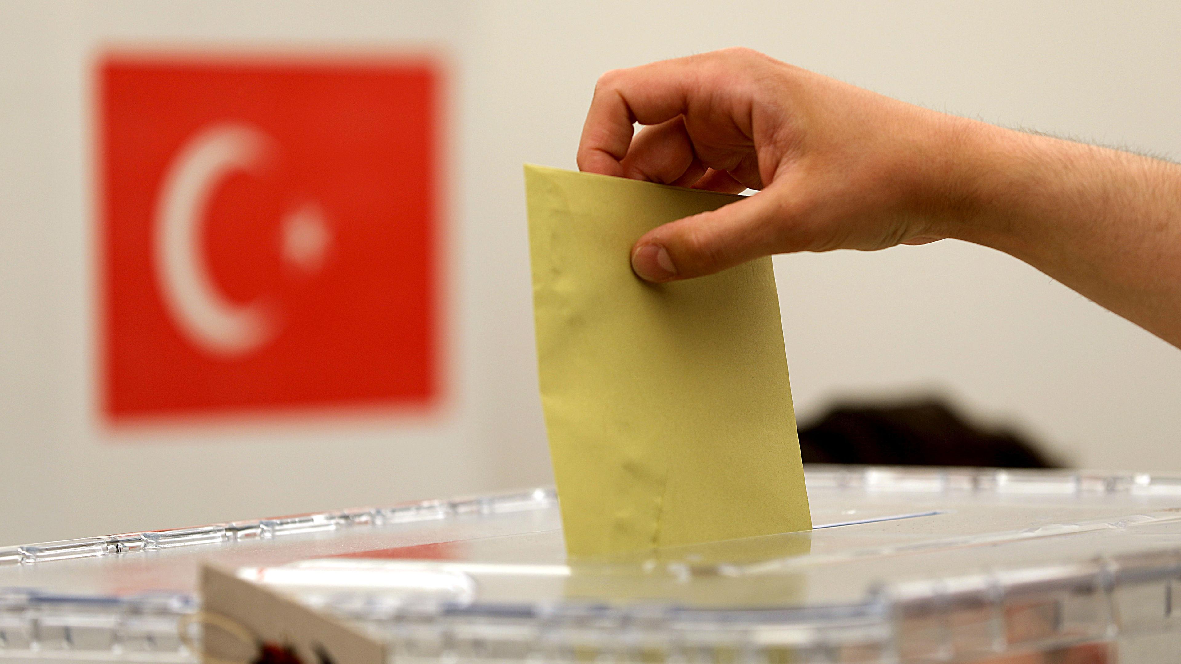 Ein Stimmzettel wird in eine Urne geworfen; im Hintergrund ist die Flagge der Türkei zu sehen.