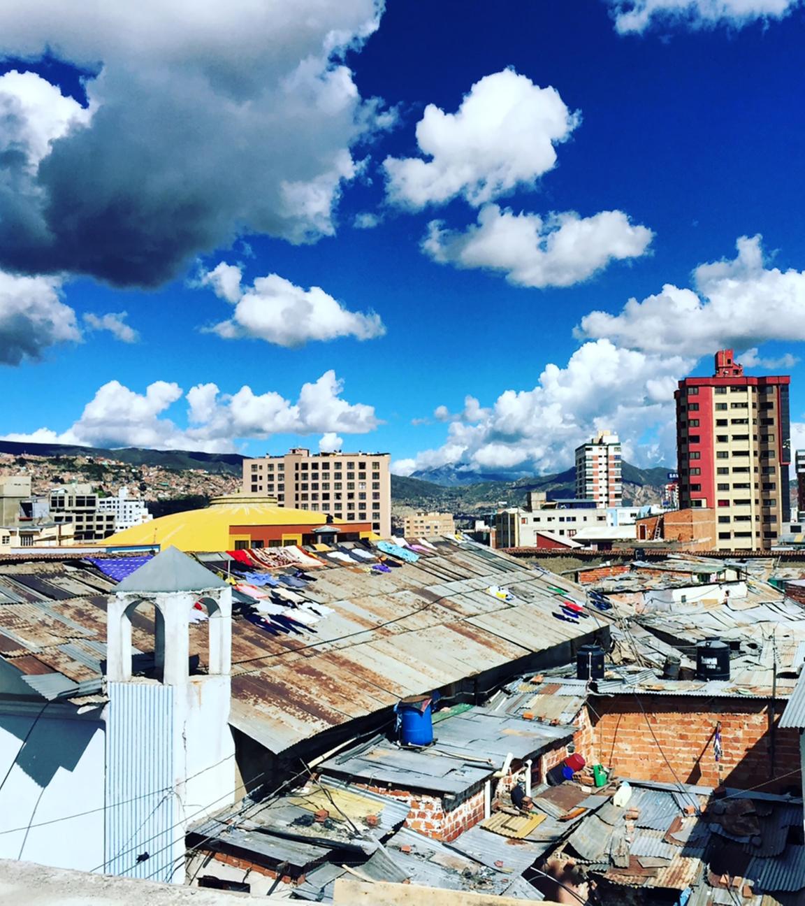 Blick über La Paz: Heruntergekommene Wellblechdächer liegen vor höheren Wohnblocks unter blauem Himmel mit einigen Wolken.