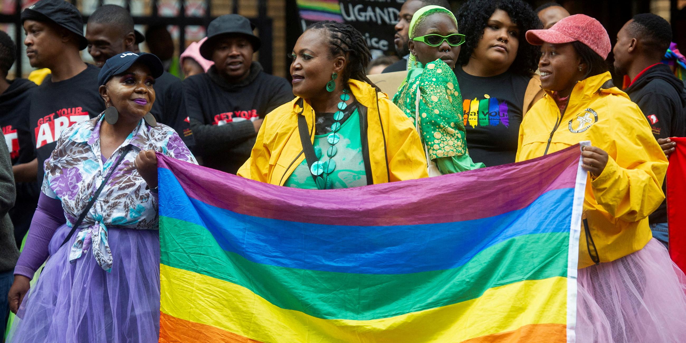 Bunt gekleidete Menschen auf einem Protest. Im Mittelpunkt steht eine Frau, die eine Regenbogenflagge hält.