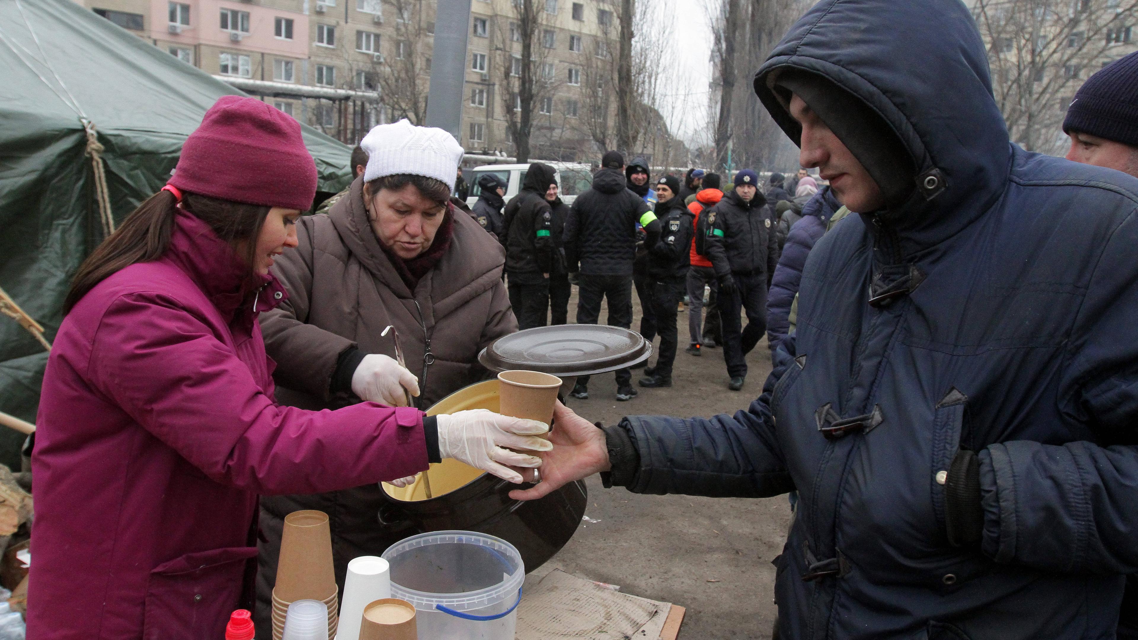 Menschen bieten Speisen und Getränke vor einem Wohnblock an, der von einer Rakete getroffen wurde