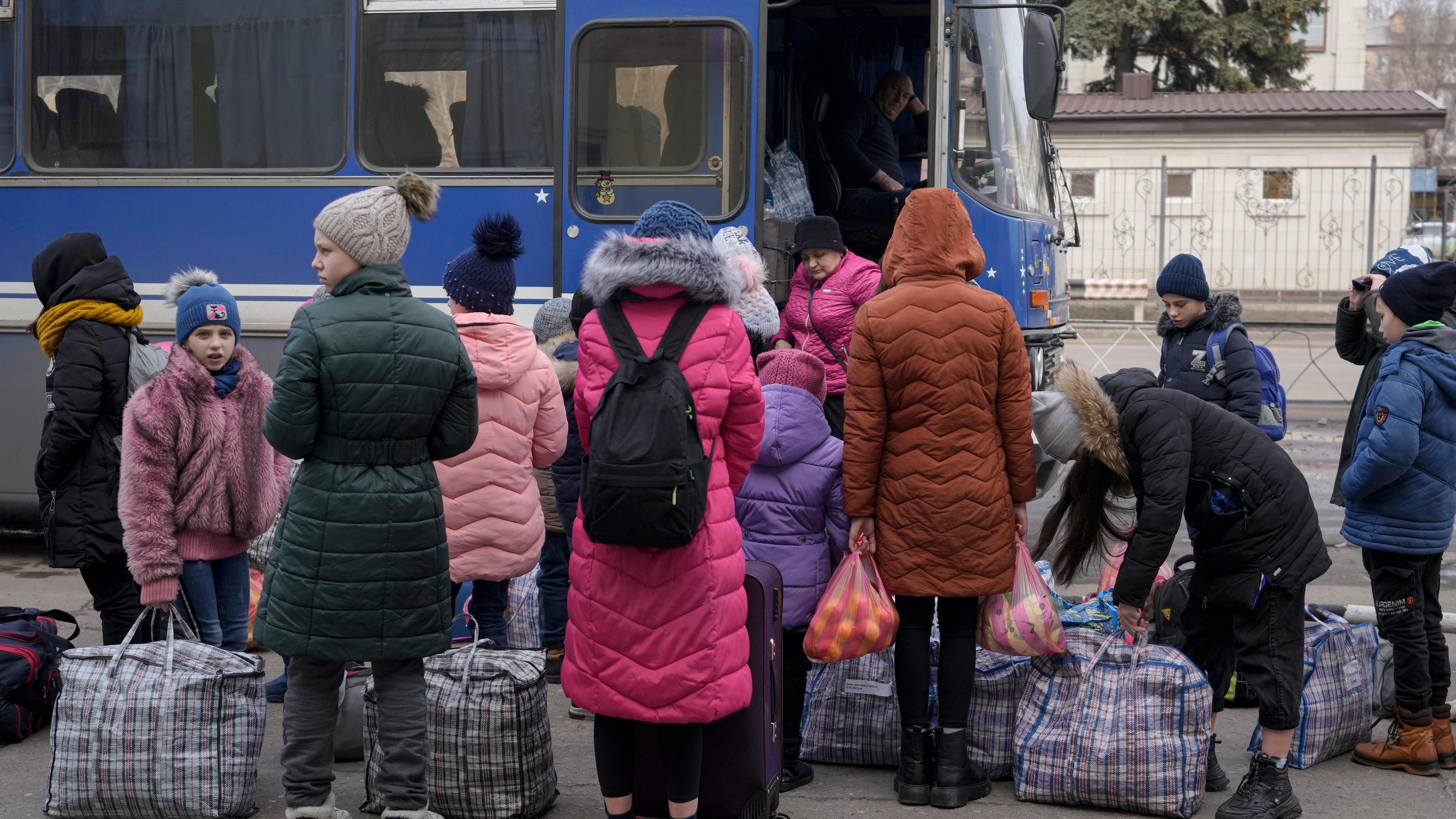 Vor einem Bus warten ukrainische Kinder mit großen Taschen, dass sie einsteigen können.