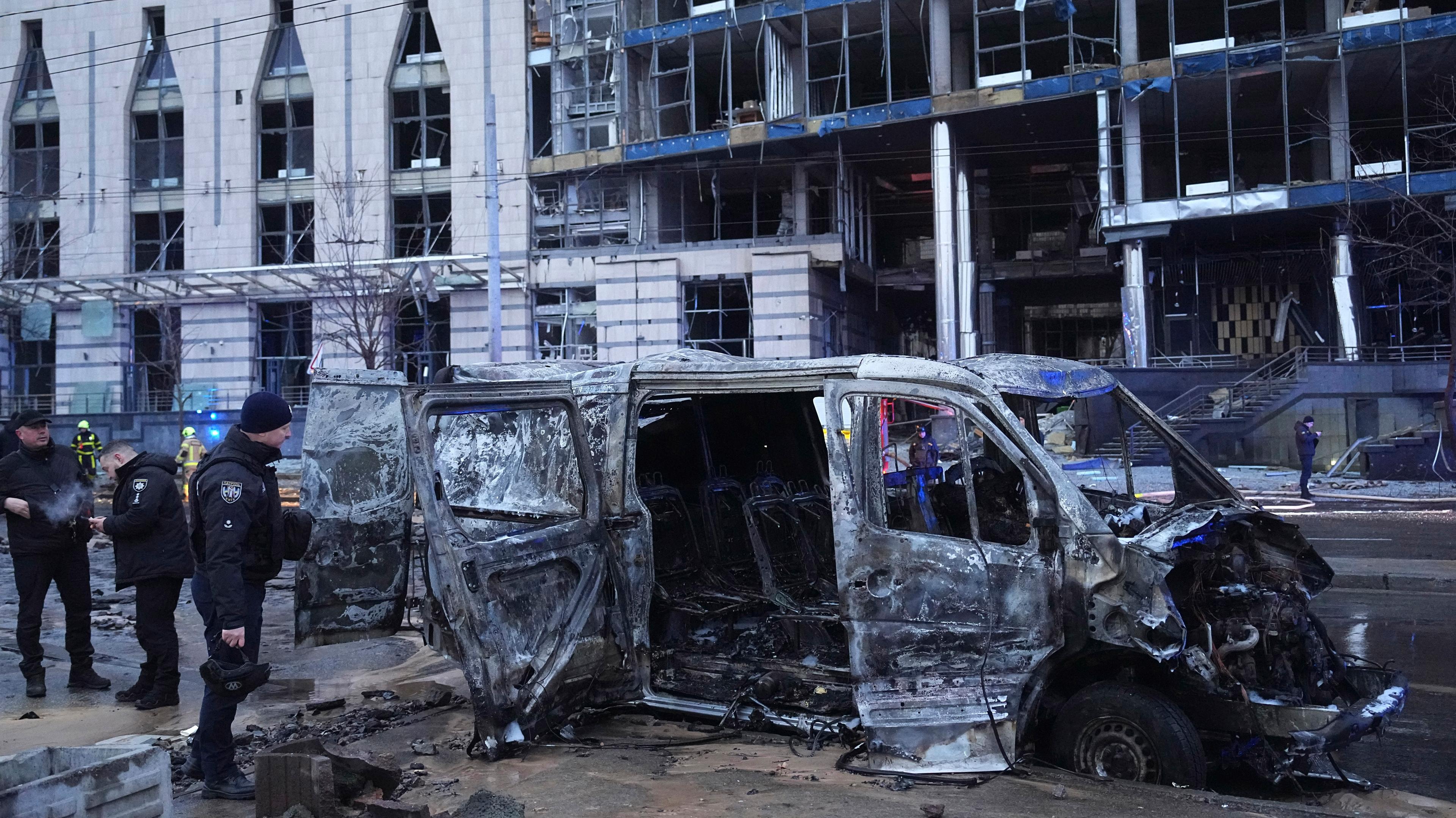 Ein zerstörtes Auto ist zu sehen, während Polizisten vor einem beschädigten Gebäude nach einem russischen Raketenangriff arbeiten.