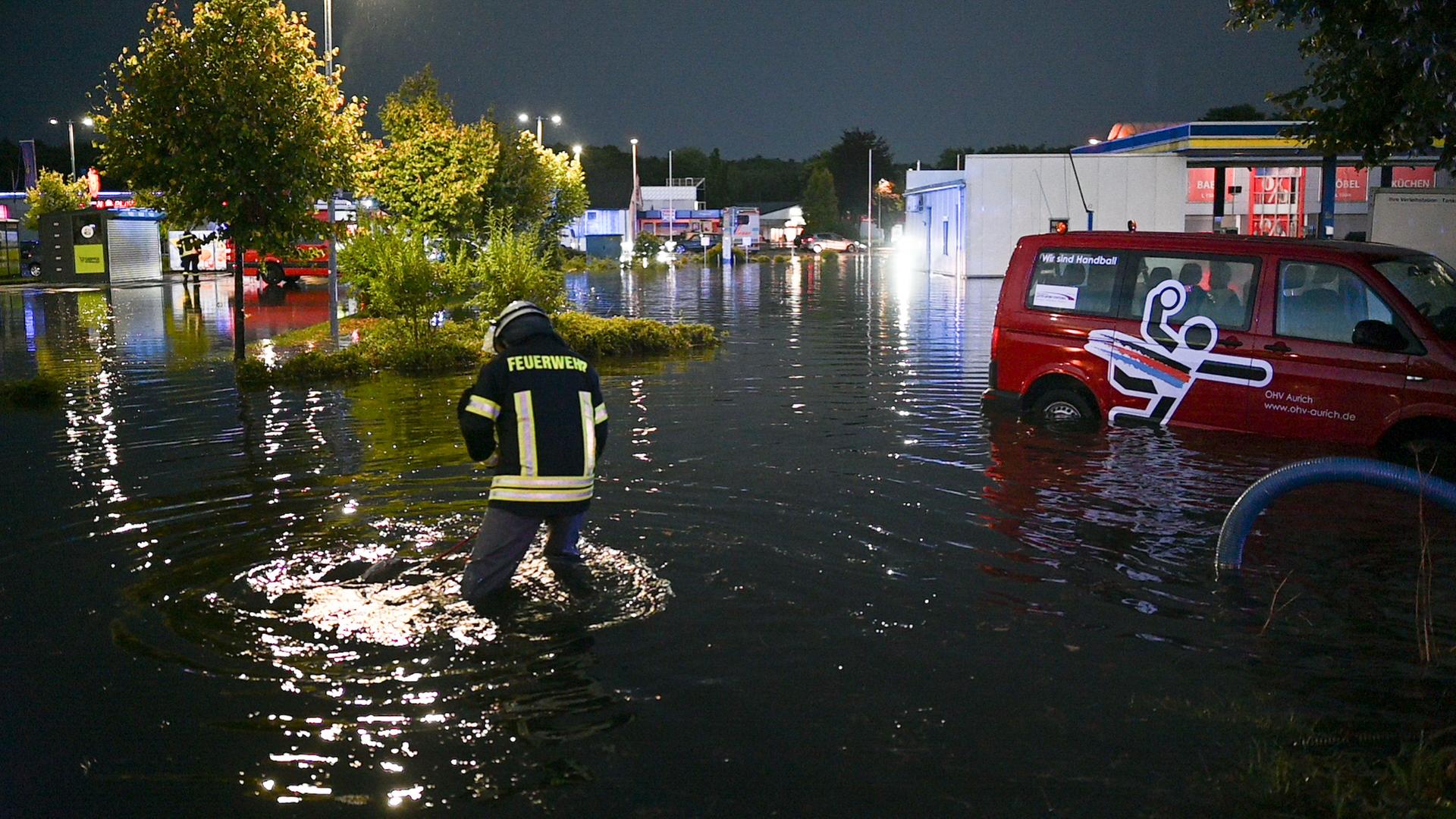 Ein Feuerwehrmann watet durch kniehohes Wasser. Neben ihm steht ein Auto im Wasser. Im Hintergrund sind eine Tankstelle und Bäume zu sehen. 