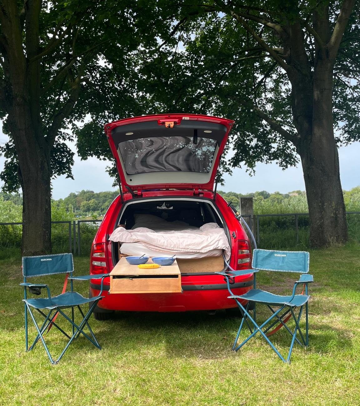 Ein rotes Auto steht mit offenem Kofferraum auf einer Wiese. Innen sieht man eine Matratze. Vor dem Auto stehen zwei Campingstühle.