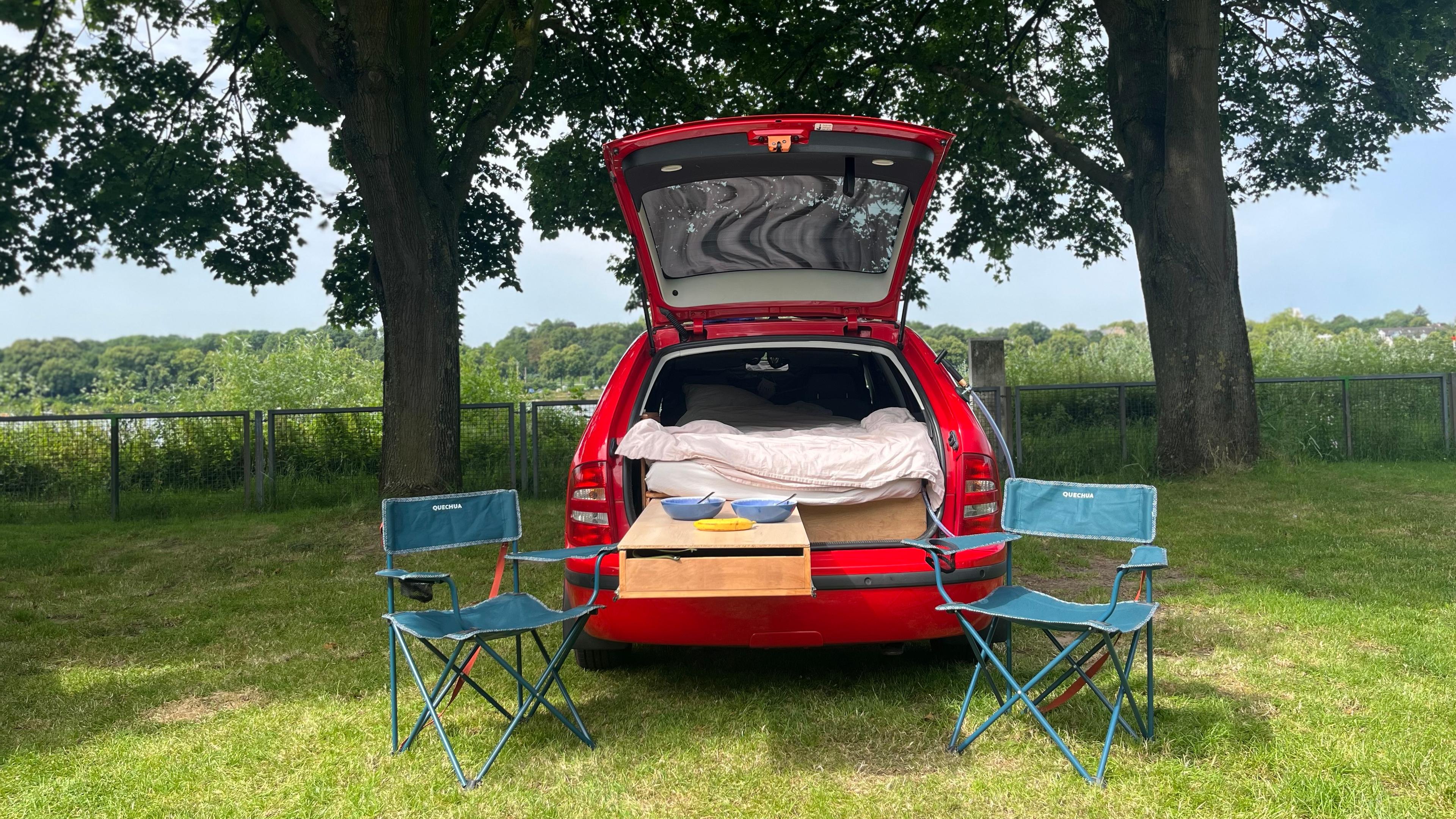 Ein rotes Auto steht mit offenem Kofferraum auf einer Wiese. Innen sieht man eine Matratze. Vor dem Auto stehen zwei Campingstühle.